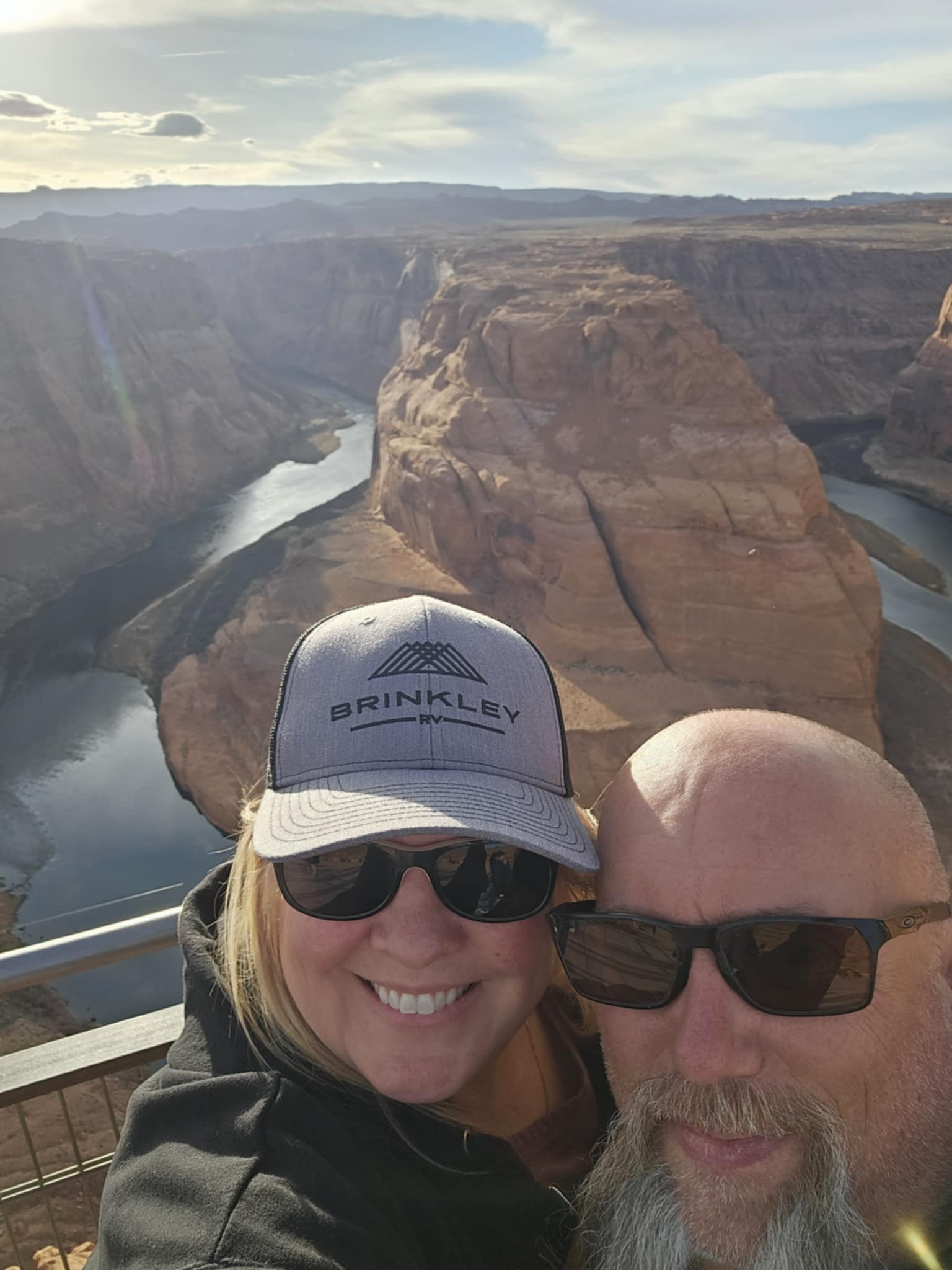 Joe and Tina selfie at Horseshoe Bend overlook with the Colorado River canyon behind them, Tina wearing a Brinkley RV cap