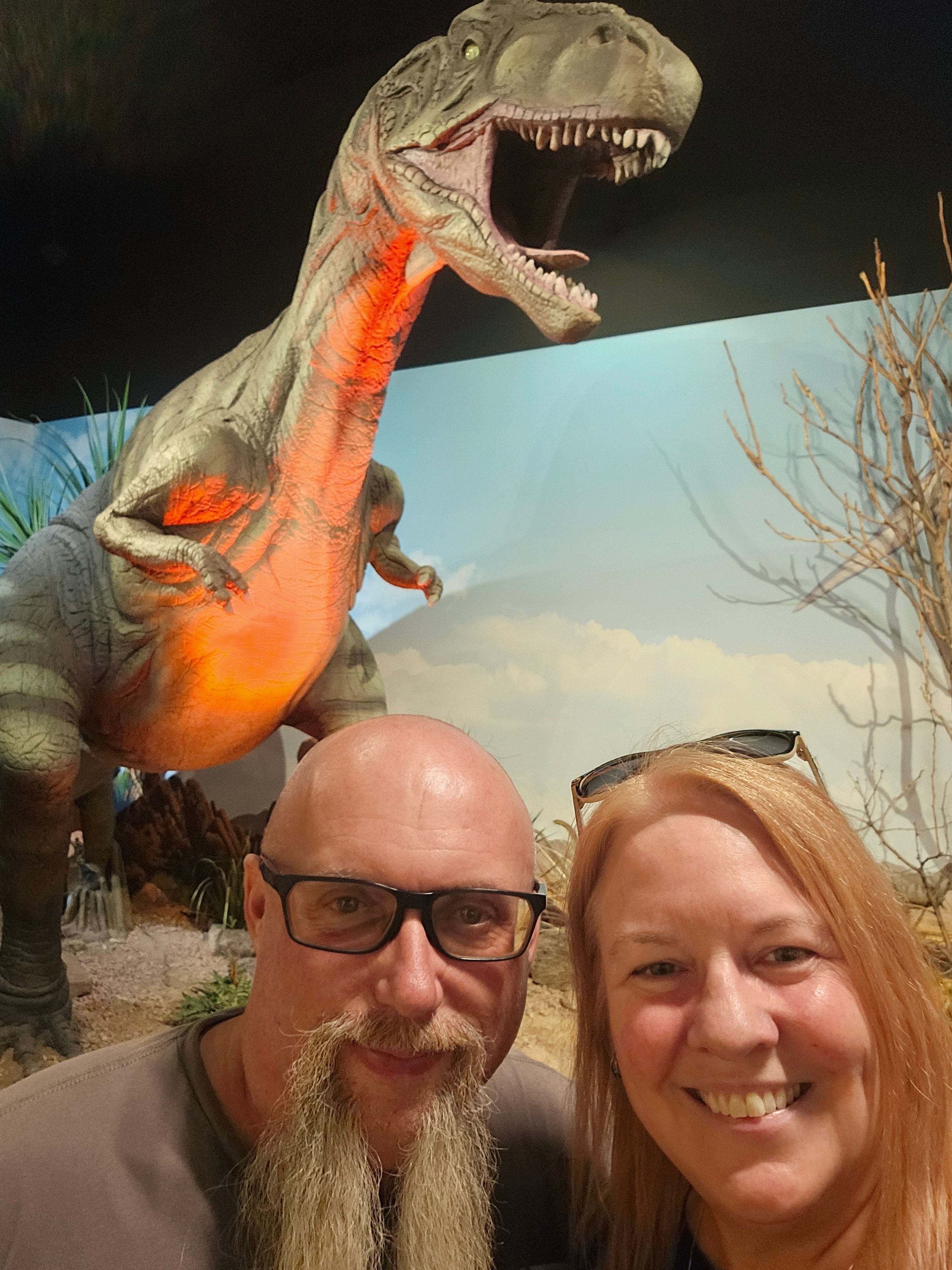 Joe and Tina selfie at The Thing museum with a roaring orange-lit T-Rex animatronic looming behind them — LOVING LIFE HAVING FUN Brinkley Z3400