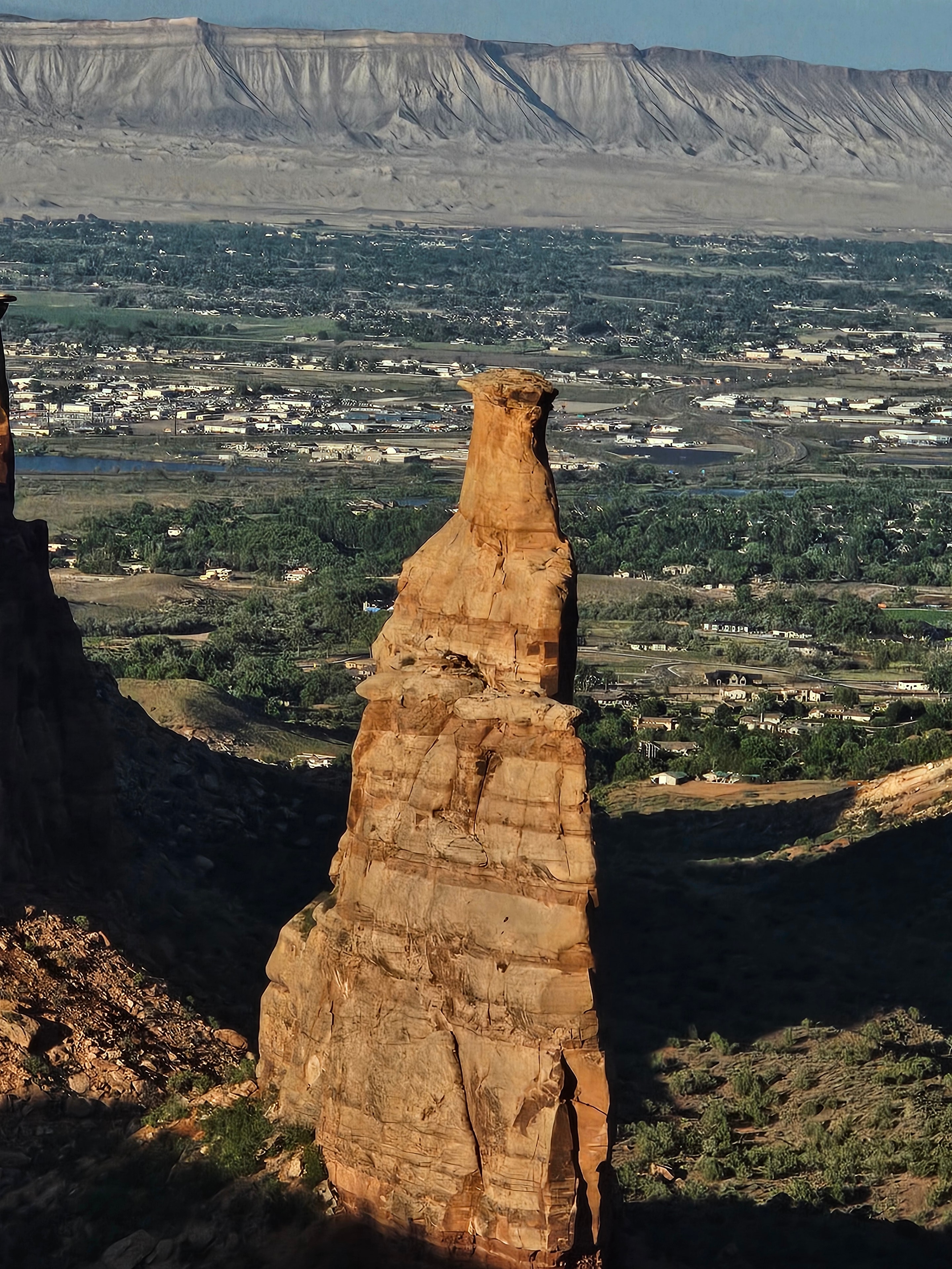 Independence Monument sandstone spire at Colorado National Monument — tall golden pillar with Grand Valley, Grand Junction, and Rocky Mountains behind in evening light — LOVING LIFE HAVING FUN Brinkley Z3400
