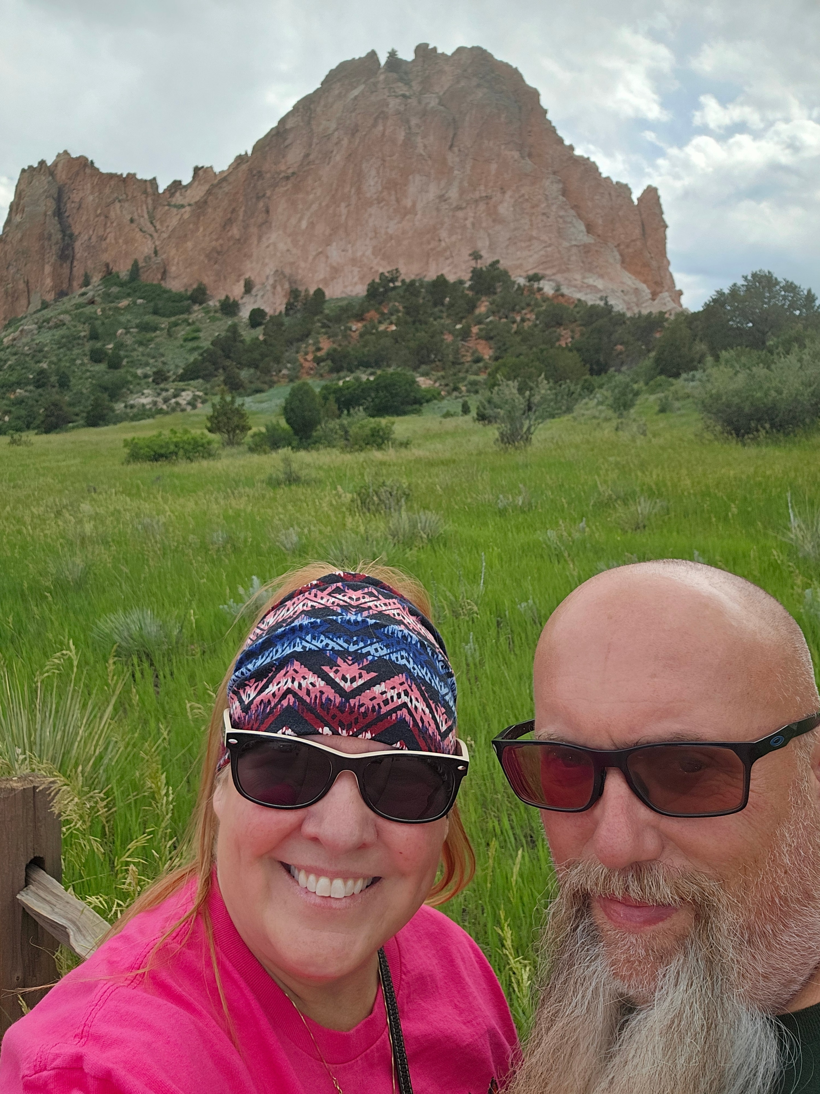 Joe and Tina selfie at Garden of the Gods — massive red sandstone formation rising behind, lush green meadow, overcast sky — LOVING LIFE HAVING FUN Brinkley Z3400