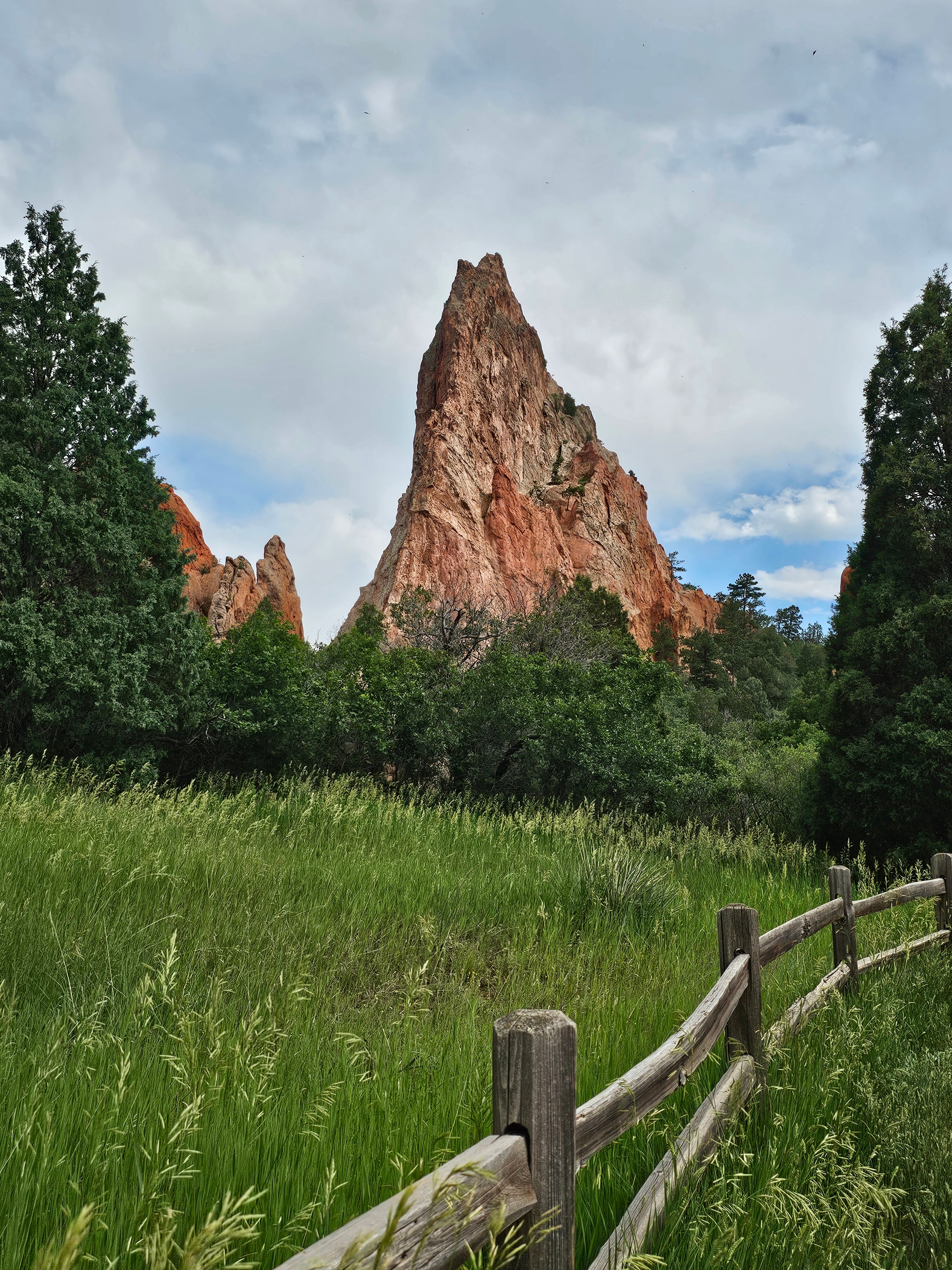 Twin red sandstone spires at Garden of the Gods — split-rail fence foreground, lush green meadow, overcast sky — LOVING LIFE HAVING FUN Brinkley Z3400