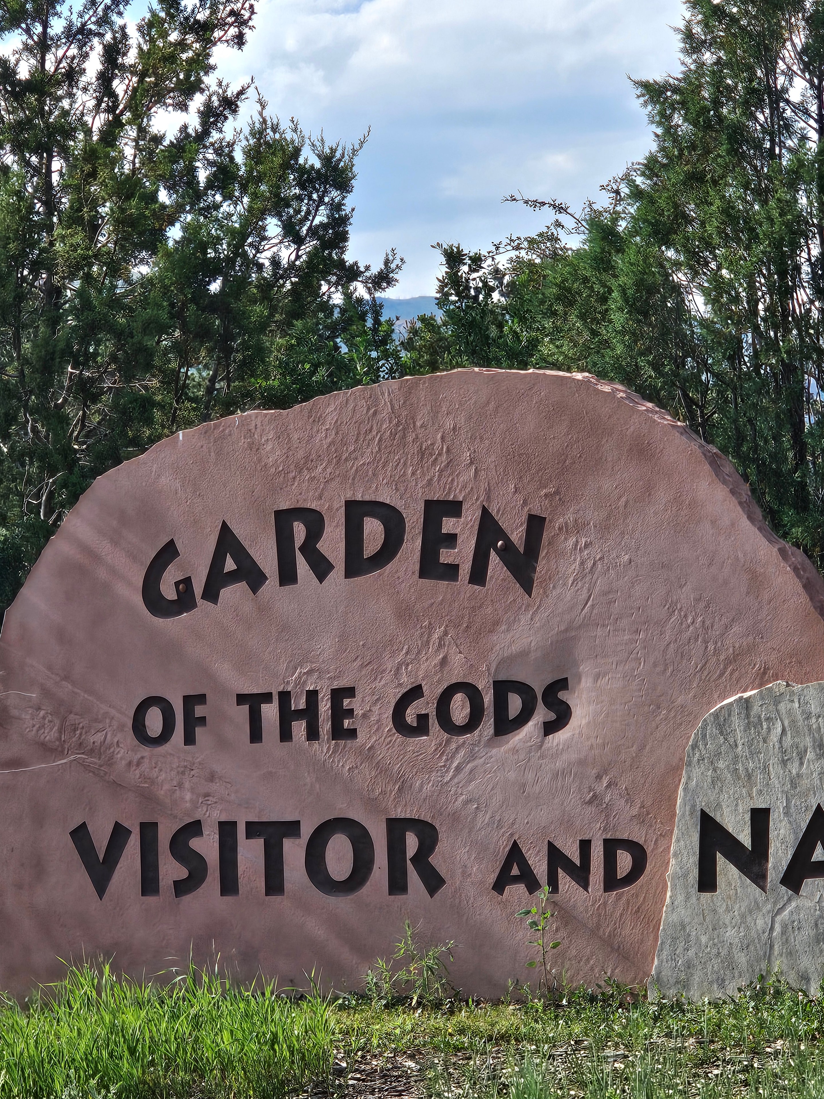 Garden of the Gods Visitor and Nature Center entrance sign — large carved sandstone boulder with text, green pines and blue sky — LOVING LIFE HAVING FUN Brinkley Z3400