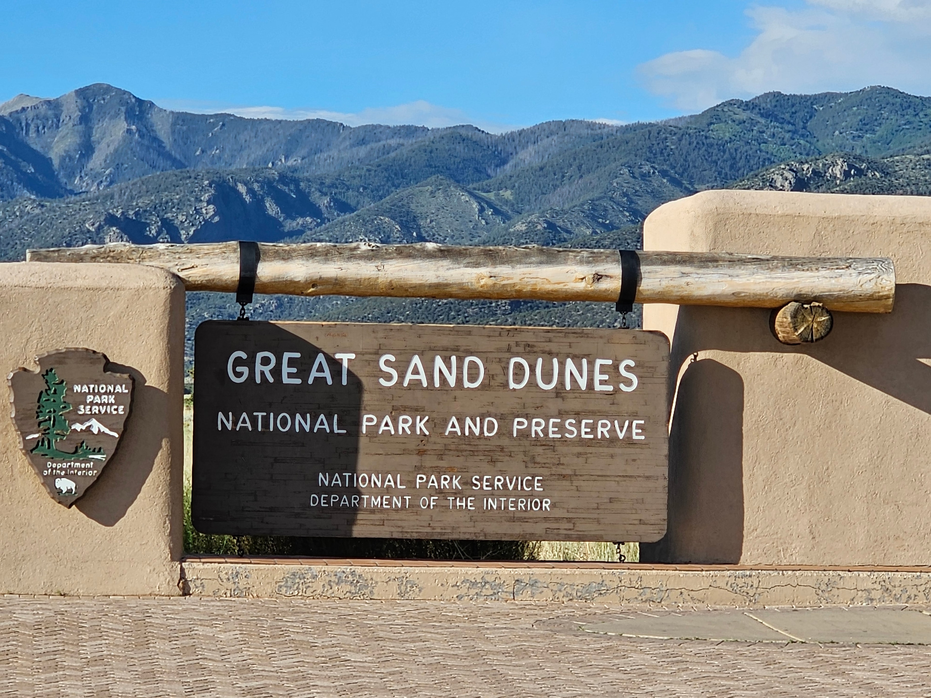 Great Sand Dunes National Park and Preserve entrance sign — adobe-style gate with log crossbeam, NPS arrowhead, Sangre de Cristo Mountains behind — LOVING LIFE HAVING FUN Brinkley Z3400