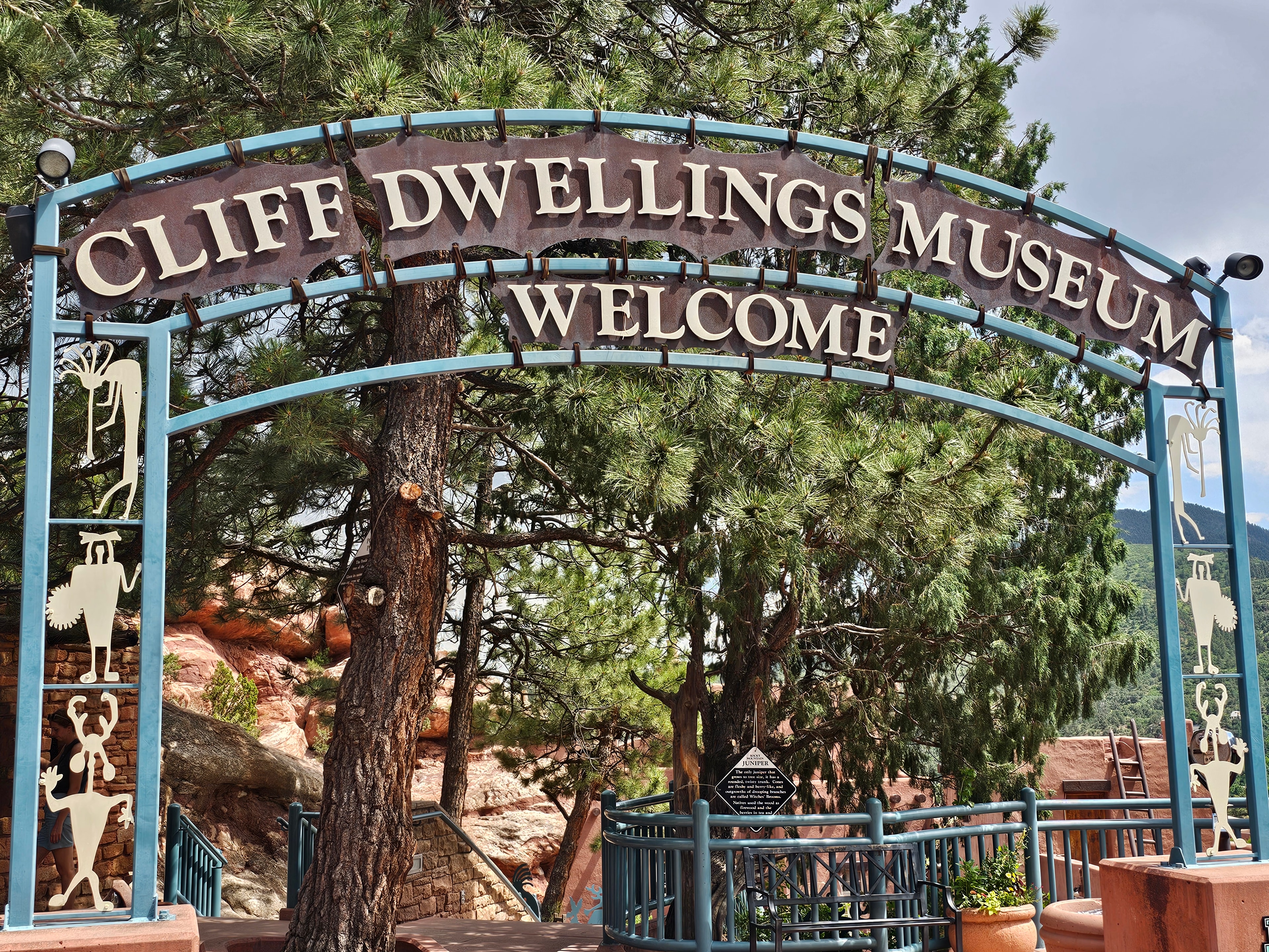 Manitou Cliff Dwellings Museum welcome sign — turquoise arch with Ancestral Puebloan figures, pine trees, red sandstone behind — LOVING LIFE HAVING FUN Brinkley Z3400