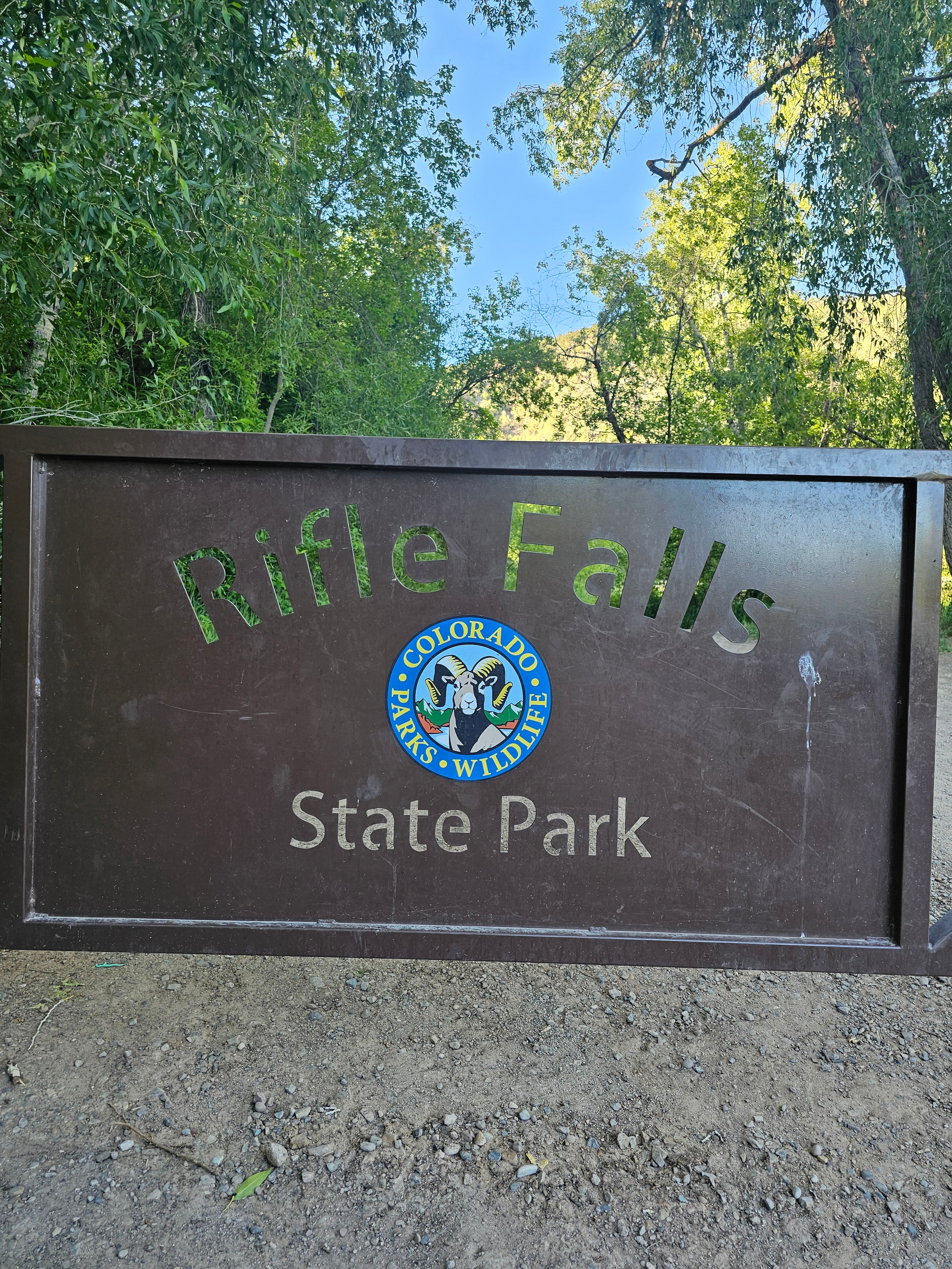 Rifle Falls State Park entrance sign — brown Colorado Wildlife Parks sign with logo, green trees behind — LOVING LIFE HAVING FUN Brinkley Z3400