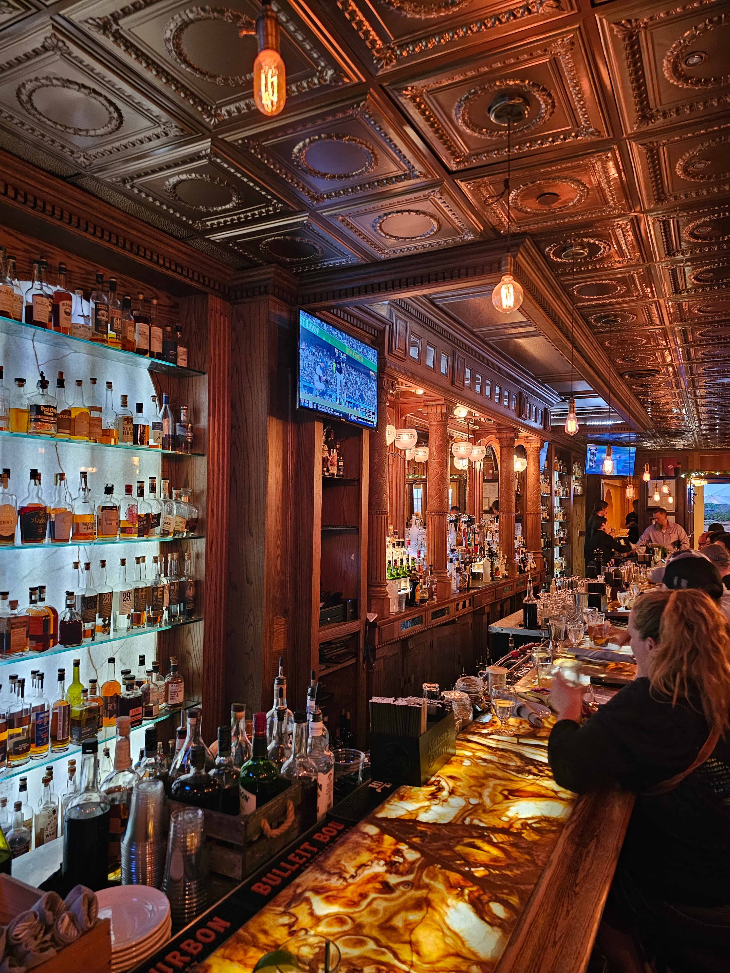 The Stanley Hotel bar interior wide — ornate copper ceiling, full bar, Bulleit Bourbon sign, warm amber glow — LOVING LIFE HAVING FUN Brinkley Z3400
