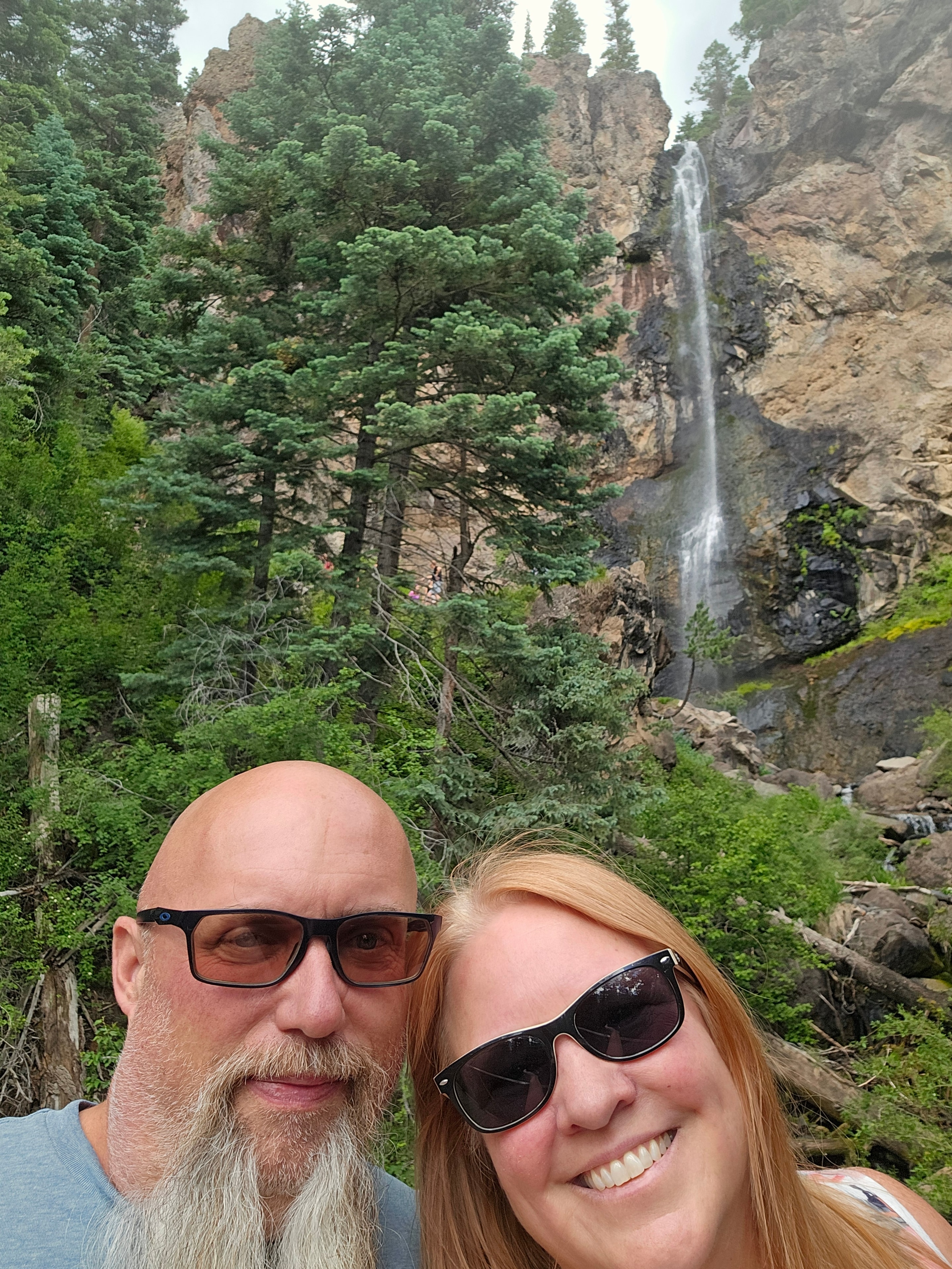 Joe and Tina selfie at Treasure Falls — waterfall cascading behind them, lush green cliff, Wolf Creek Pass, Colorado — LOVING LIFE HAVING FUN Brinkley Z3400