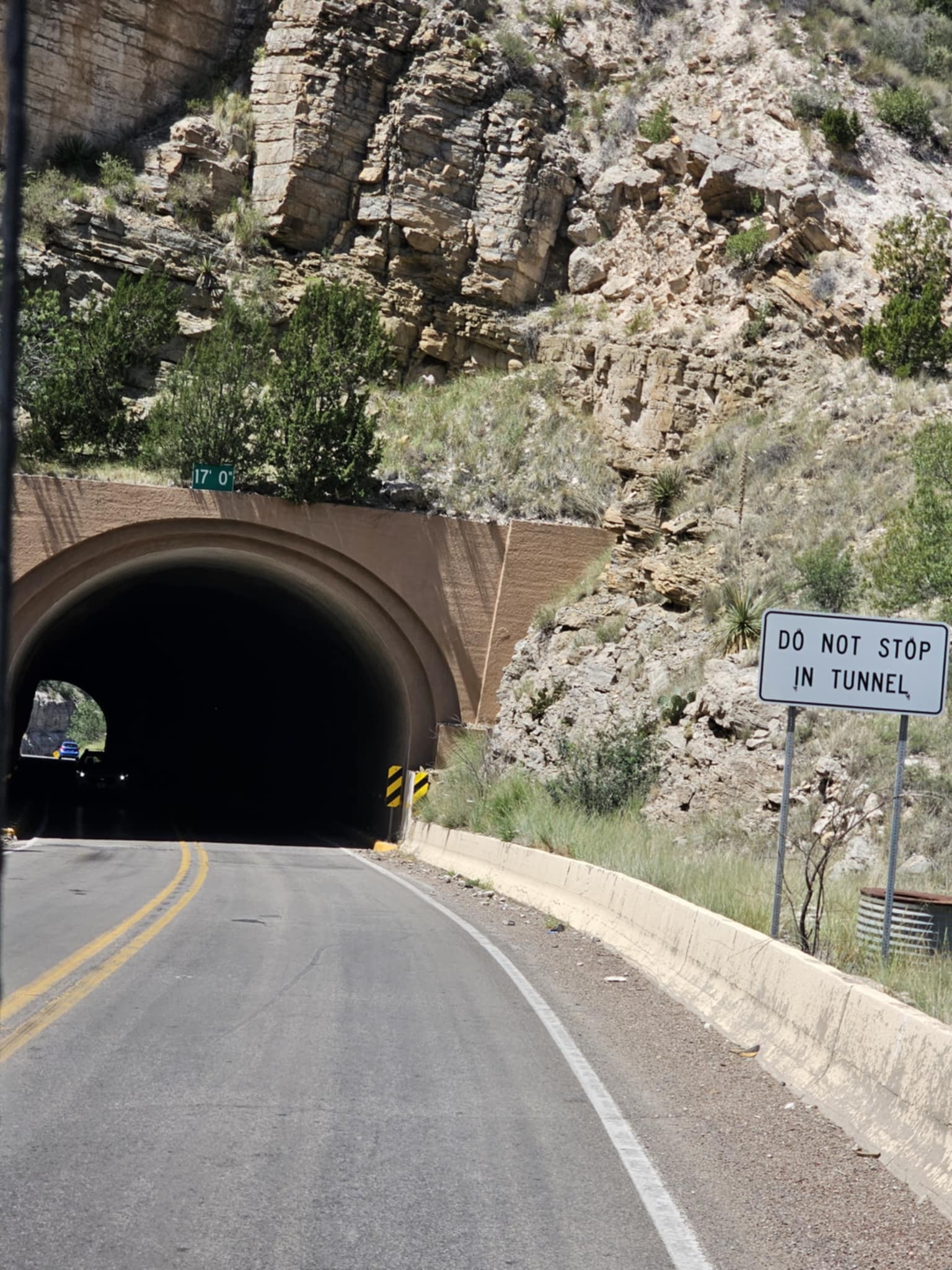 Road tunnel entrance on the drive to Alamogordo — a Do Not Stop In Tunnel sign beside the arched tunnel opening cut through tan limestone rock, yucca and desert scrub on the hillside above