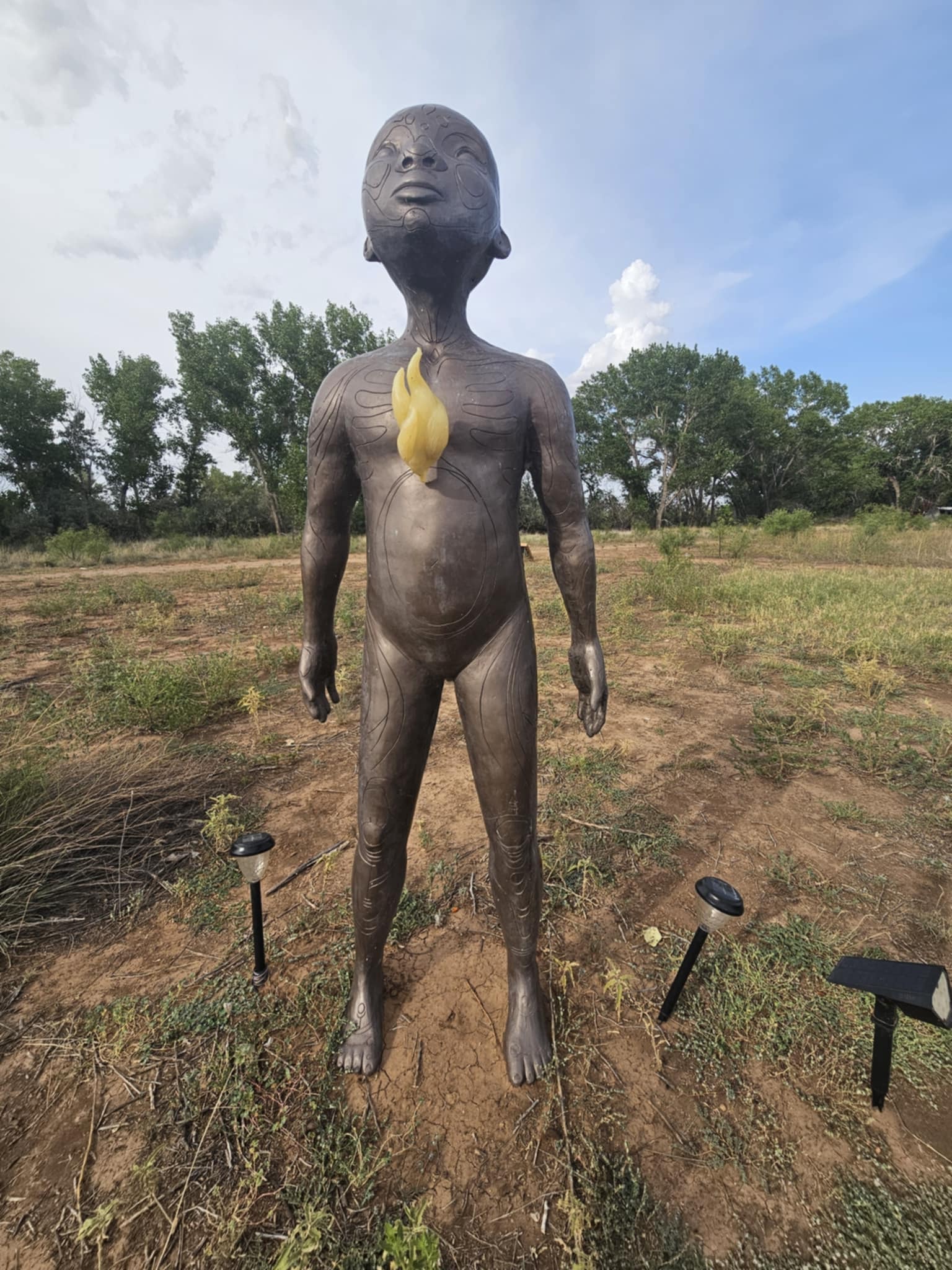 Dark patinated bronze sculpture of a child figure with intricate etched body patterns and a glowing yellow flame accent at the chest, standing in the open desert field at Art City, solar pathway lights flanking the base