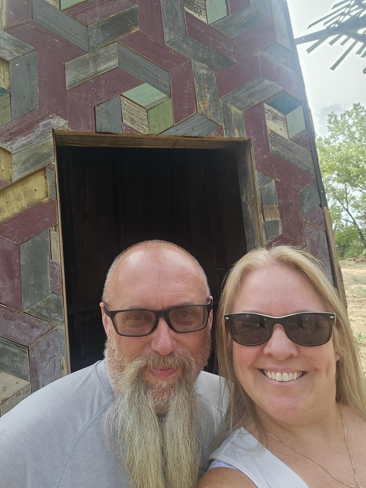 Joe and Tina selfie in front of a stunning geometric reclaimed wood art installation at Art City — the structure covered in a complex interlocking diamond pattern of weathered boards in muted greens, purples, and grays, a dark doorway opening behind them