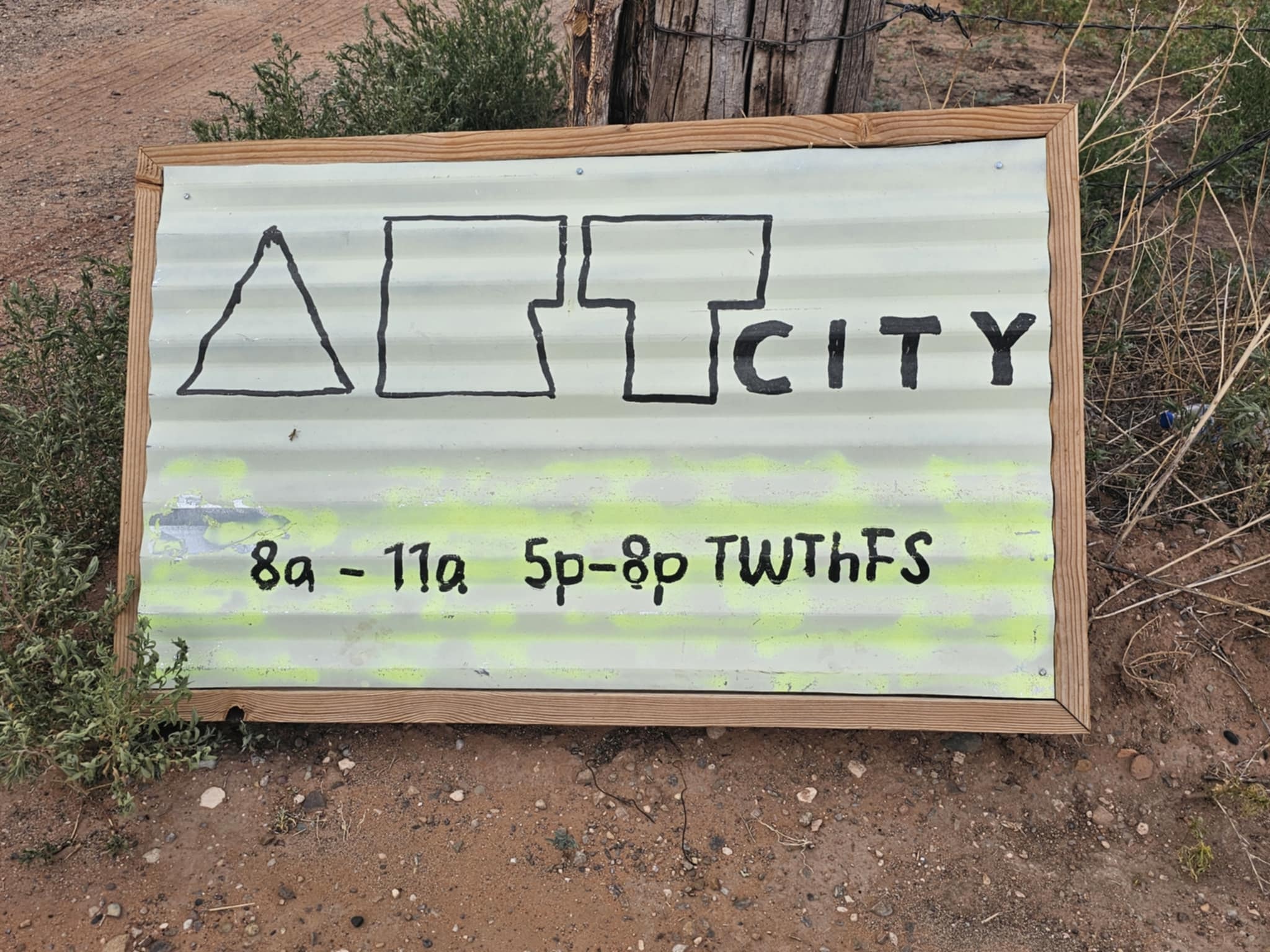 Hand-painted Art City sign on corrugated metal in a wood frame leaning against a fence post in the New Mexico desert — geometric logo of a triangle, rectangles, and house icon spelling out "Art City" with hours 8a–11a and 5p–8p Tuesday through Saturday painted in neon yellow highlight below
