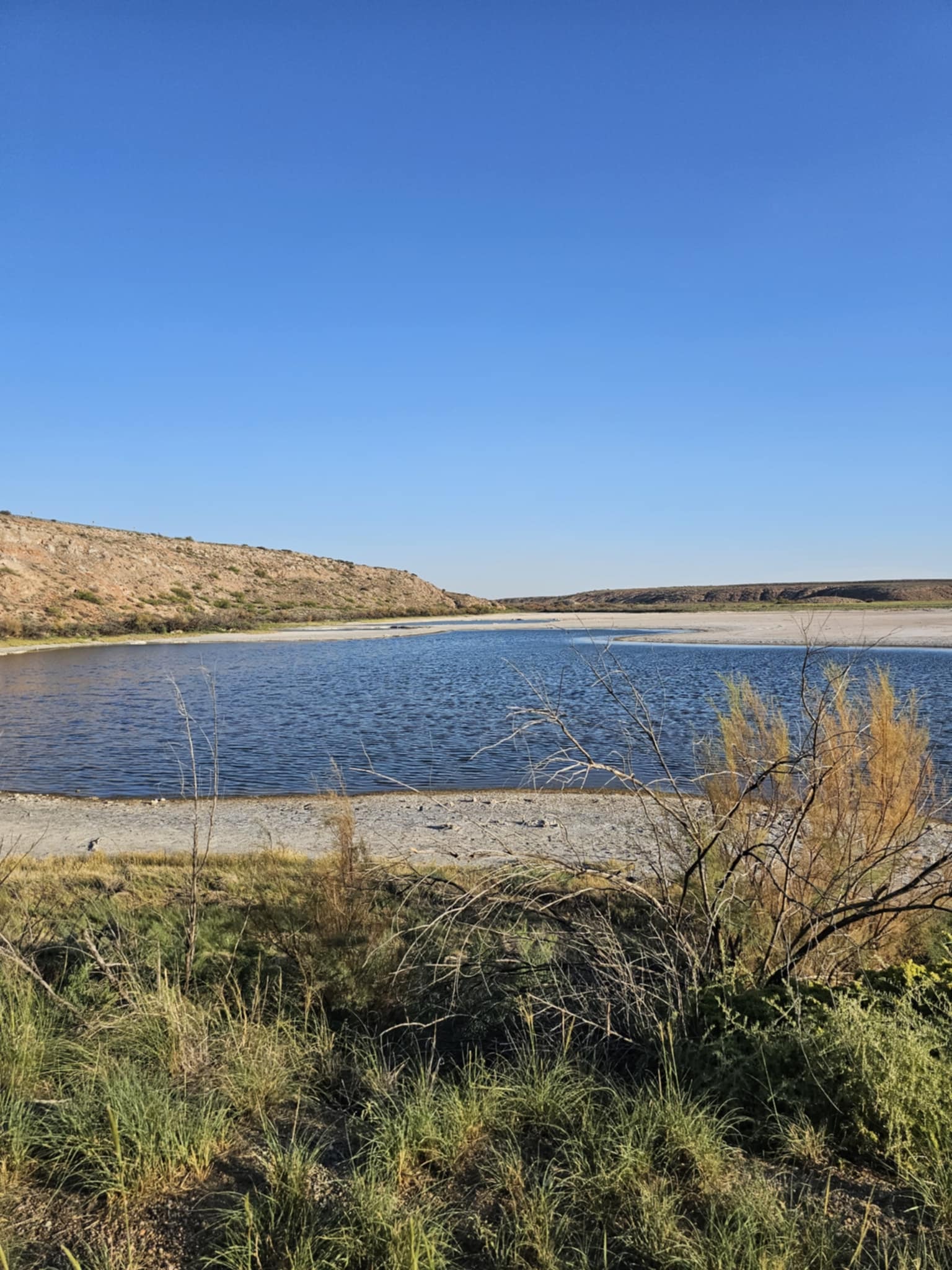 Calm blue lake at Bottomless Lakes State Park with a sandy white shore, red and tan bluffs rising on the left, golden grasses in the foreground, deep blue sky