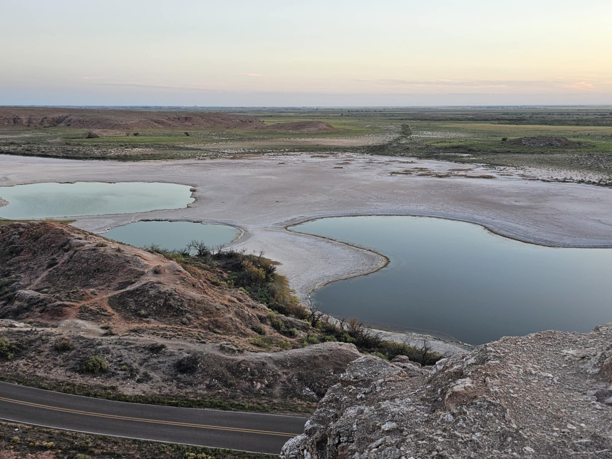Aerial overlook of Bottomless Lakes State Park at dusk — multiple vivid blue-green sinkhole pools scattered across a white salt flat, red bluffs in the foreground, the flat Chihuahuan Desert stretching to the horizon under a soft pastel sunset sky