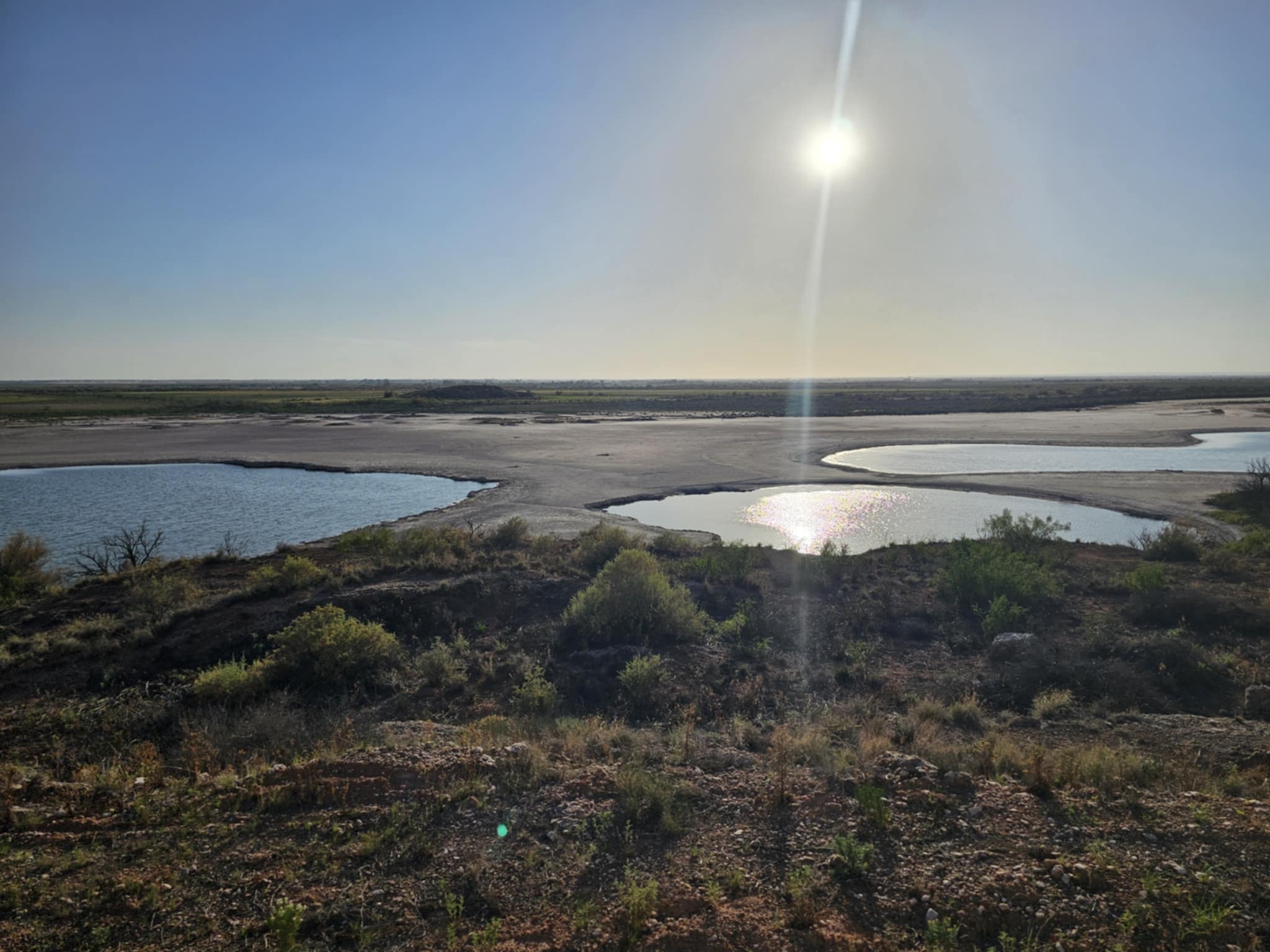 Midday overlook of the Bottomless Lakes sinkholes — multiple blue pools scattered across a white salt flat, bright sun overhead, flat desert stretching to the horizon