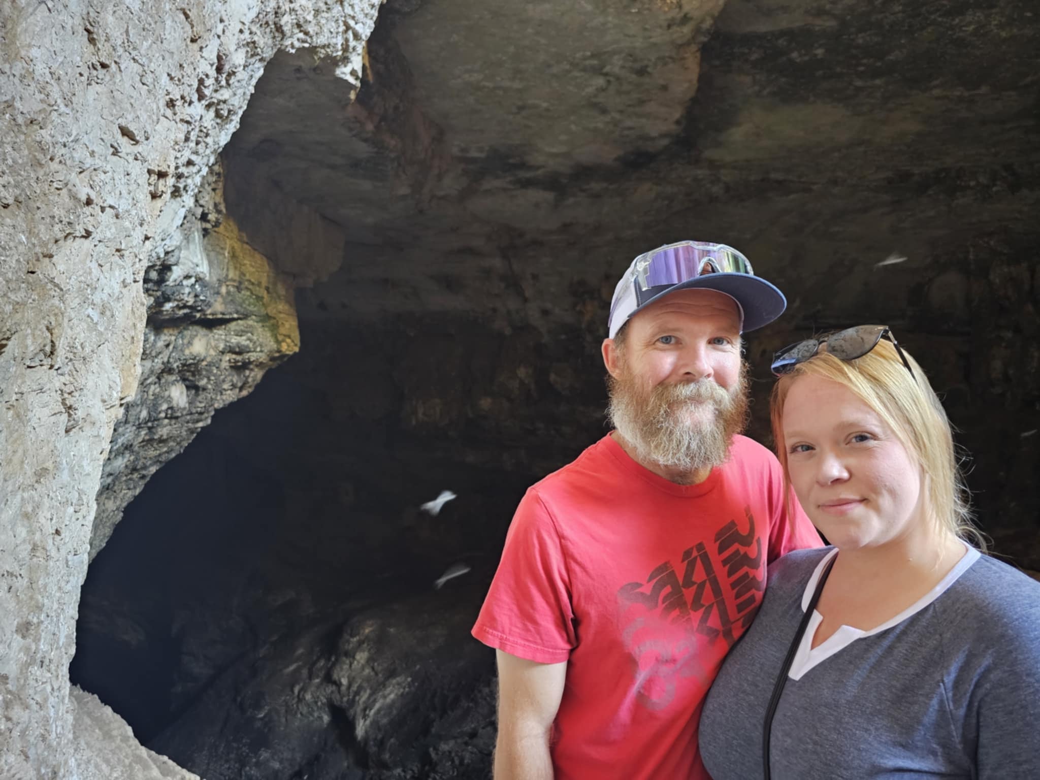 Ben and Shelby at the natural entrance of Carlsbad Caverns — standing in front of the large cave mouth opening in the tan limestone hillside, bats visible in flight in the dark passage behind them