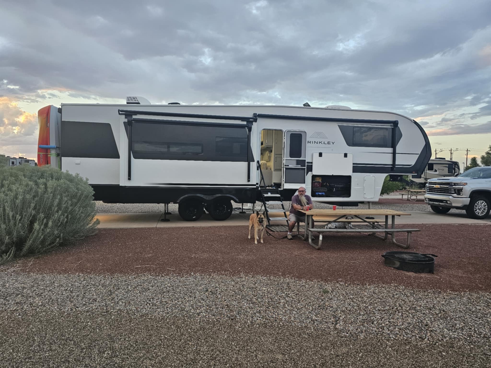 Joe sitting at the picnic table beside the Brinkley Z3400 with Zero at his side under a dramatic stormy sunset sky at the La Ventana area campsite