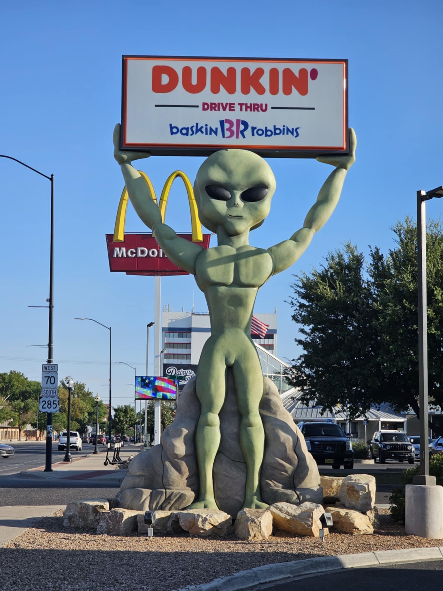 The famous green alien statue in downtown Roswell — a muscular extraterrestrial figure standing on a boulder pedestal with arms raised overhead holding a Dunkin' Drive Thru / Baskin-Robbins sign, the McDonald's golden arches and downtown Roswell visible behind it under a clear blue sky