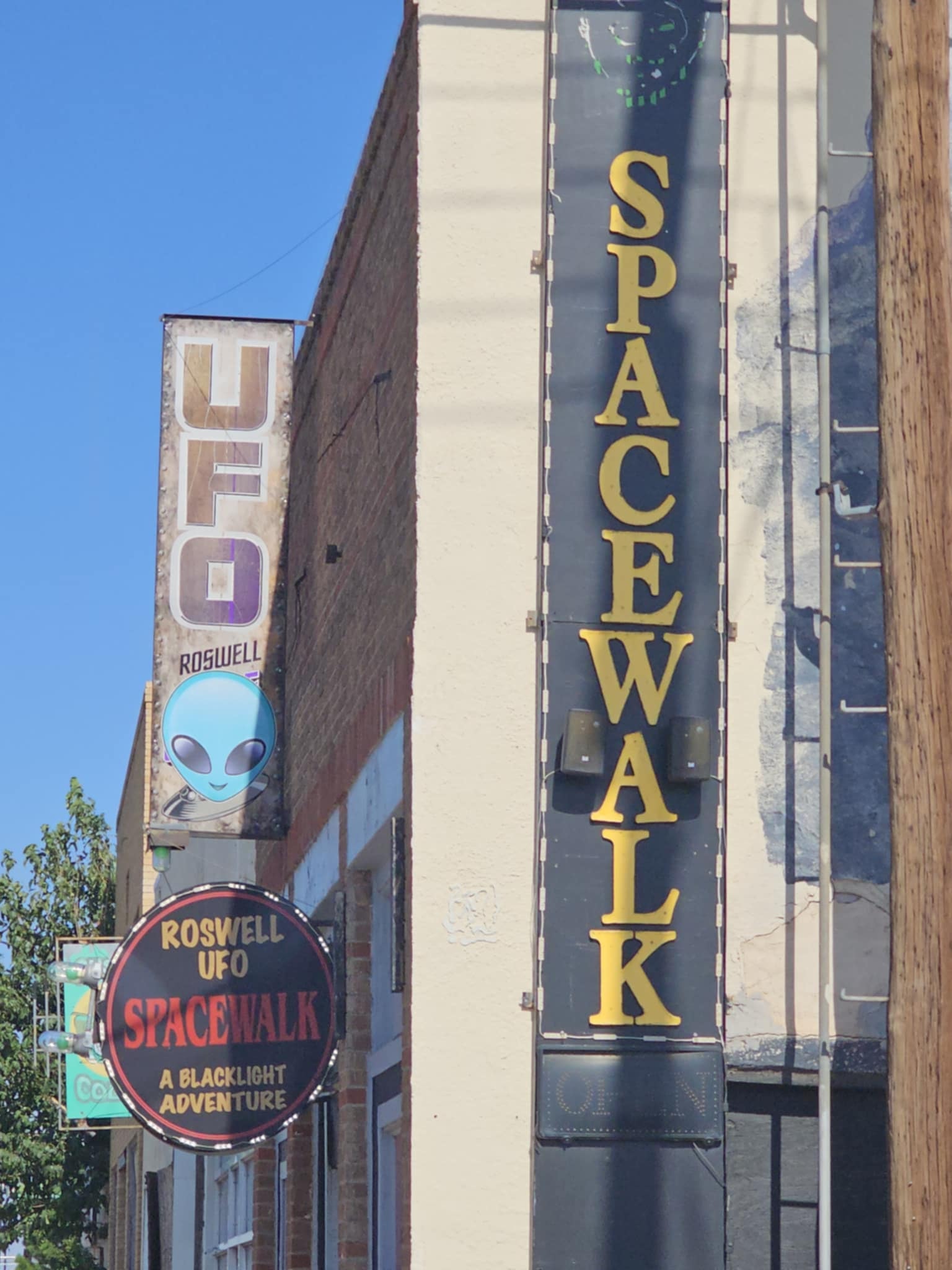 Downtown Roswell UFO-themed signage — the weathered "UFO Roswell" blade sign with a teal alien face below it, the circular "Roswell UFO Spacewalk — A Blacklight Adventure" sign, and the tall gold "SPACEWALK" letters mounted vertically on a dark building facade, all under a vivid blue New Mexico sky