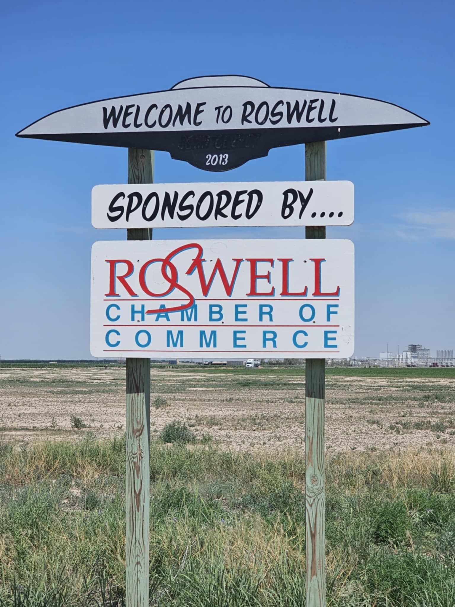 "Welcome to Roswell" roadside sign shaped like a flying saucer on two wooden posts — the UFO silhouette reads "Welcome to Roswell" with "2013" below, a second panel reads "Sponsored by.... Roswell Chamber of Commerce" in red and blue lettering, flat New Mexico farmland stretching to the horizon behind it