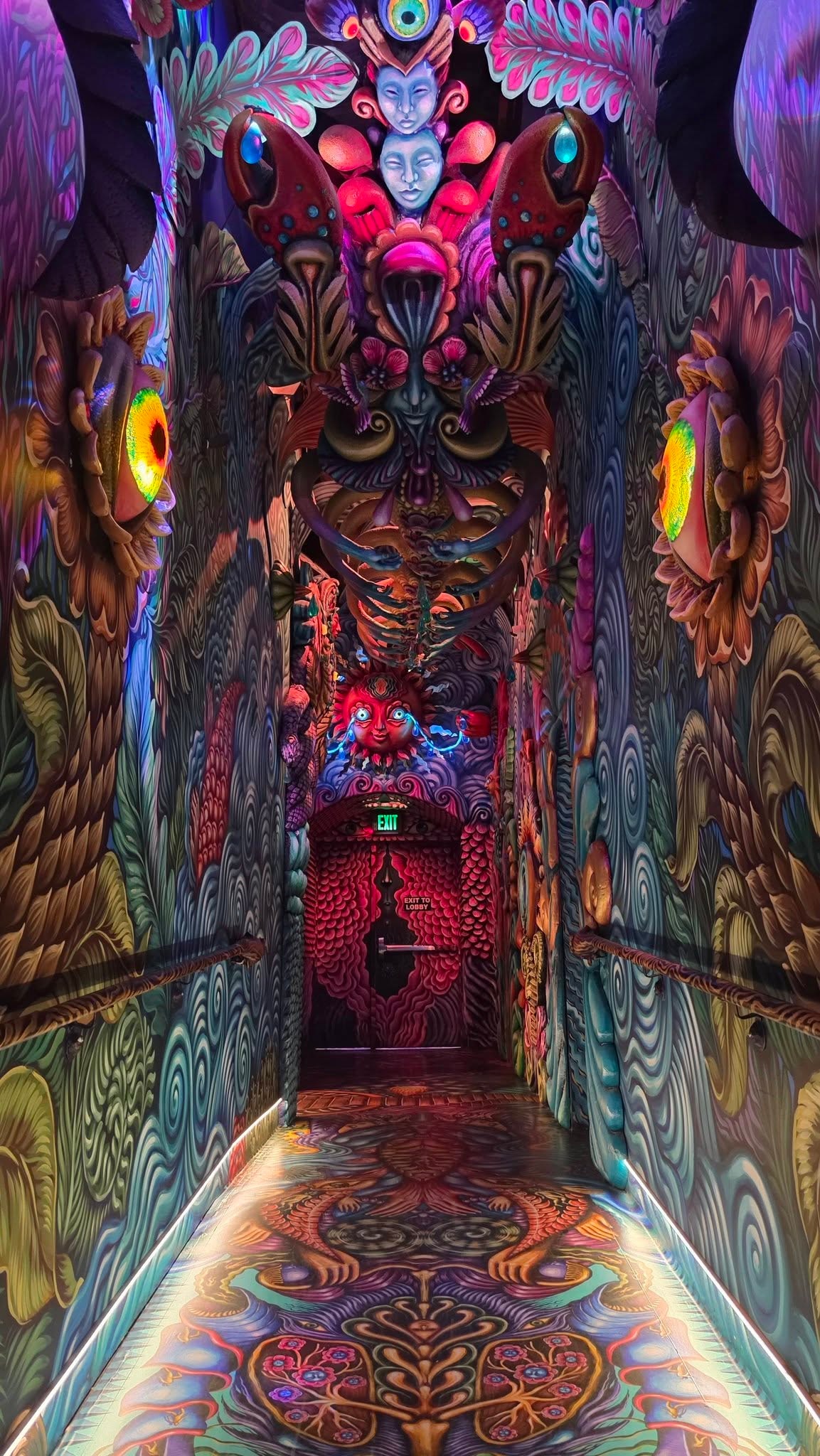 Immersive psychedelic corridor inside Meow Wolf House of Eternal Return Santa Fe — floor-to-ceiling surrealist murals in deep teal, magenta, crimson, and gold cover every surface including the floor, a demonic face with glowing eyes looms above the exit door at the far end, circular glowing orbs flank the walls