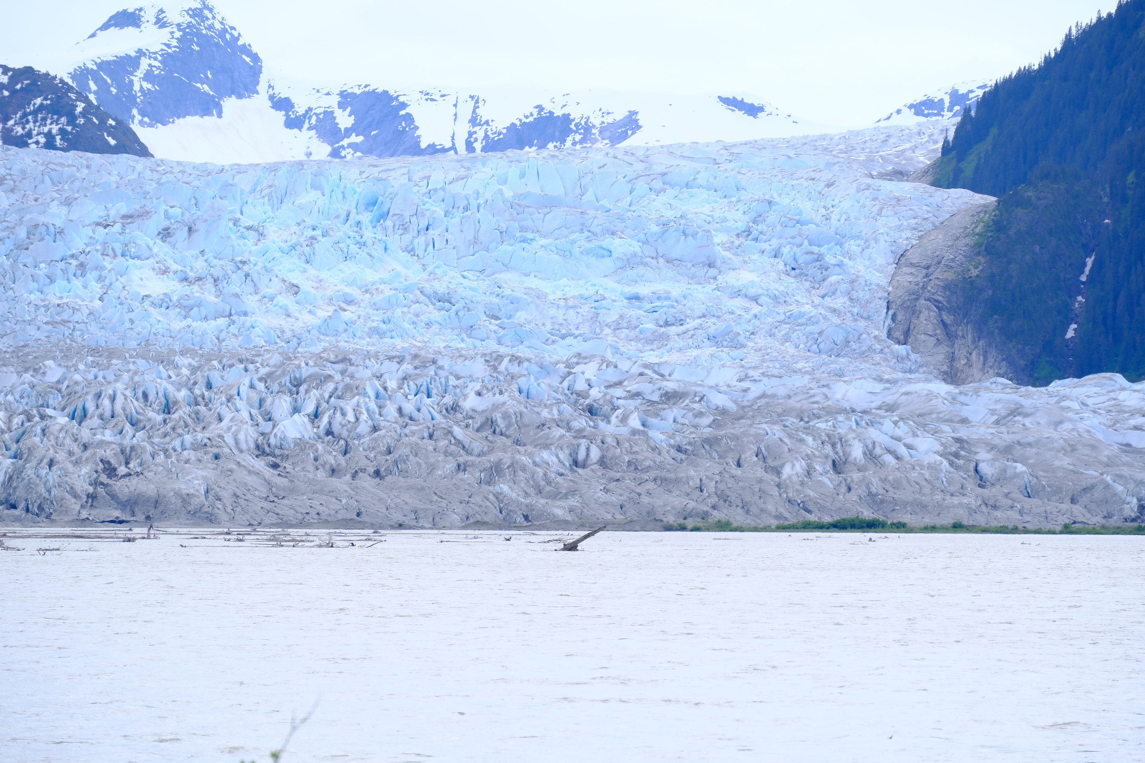 The Taku Glacier face from the lodge grounds — a vast expanse of blue-white fractured ice and seracs stretching across the full frame, the glacier terminus meeting the silty river flats