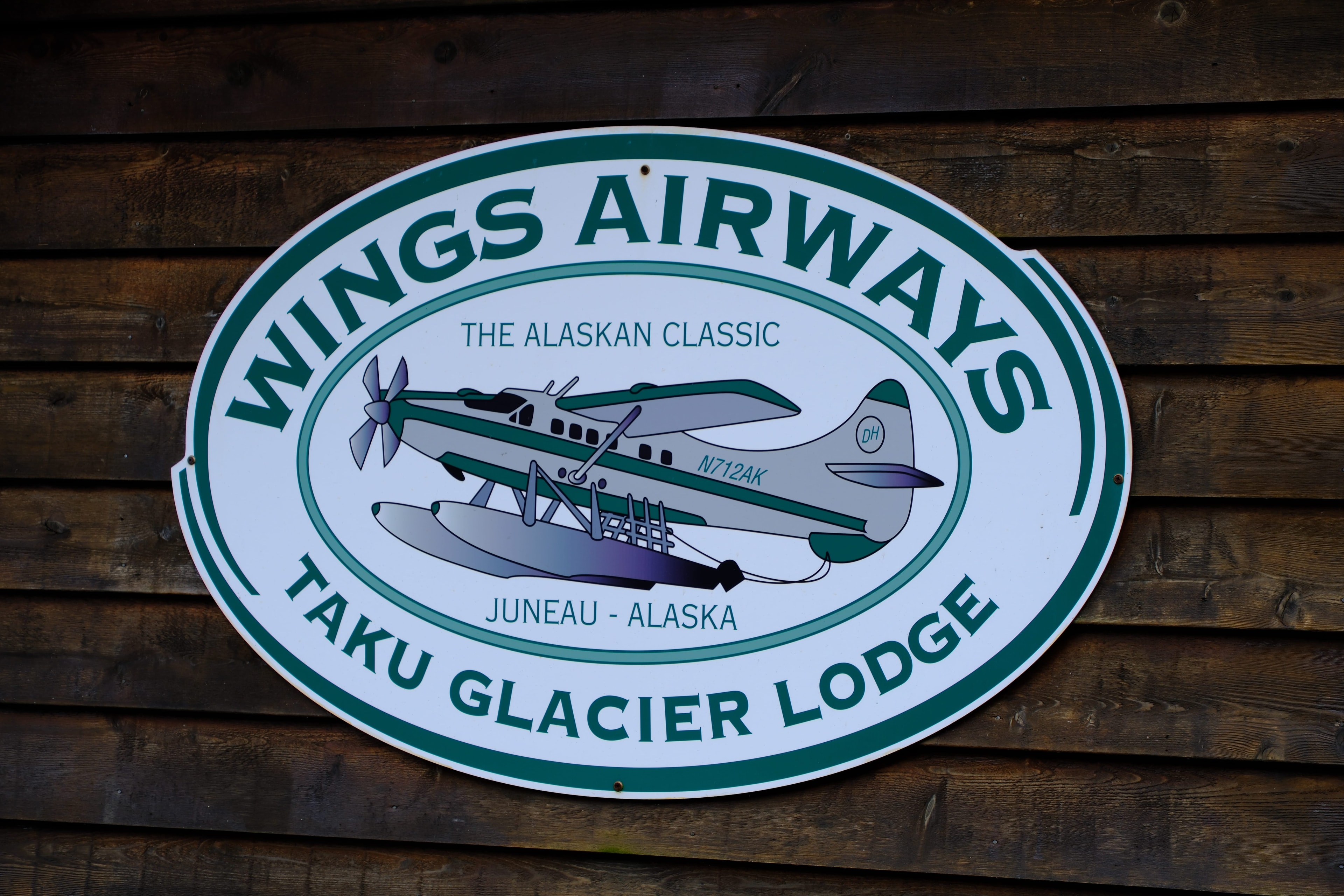 A Wings Airways floatplane lifting off the Gastineau Channel — the seaplane banking over the calm water with Juneau's waterfront and forested mountains rising behind it