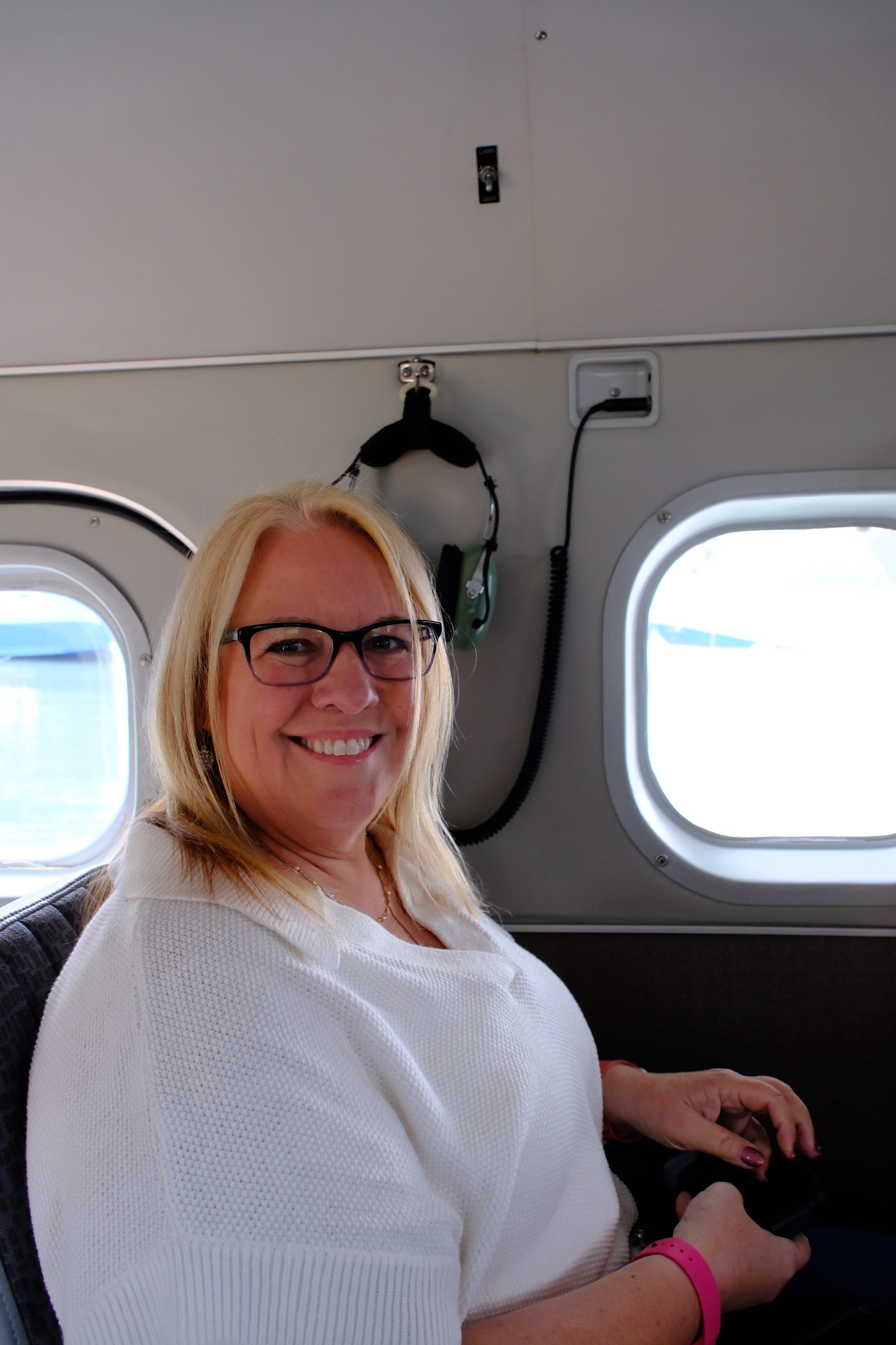 Tina smiling inside the floatplane cabin — headset around her neck, two oval windows behind her showing blue sky, pure joy on her face before takeoff