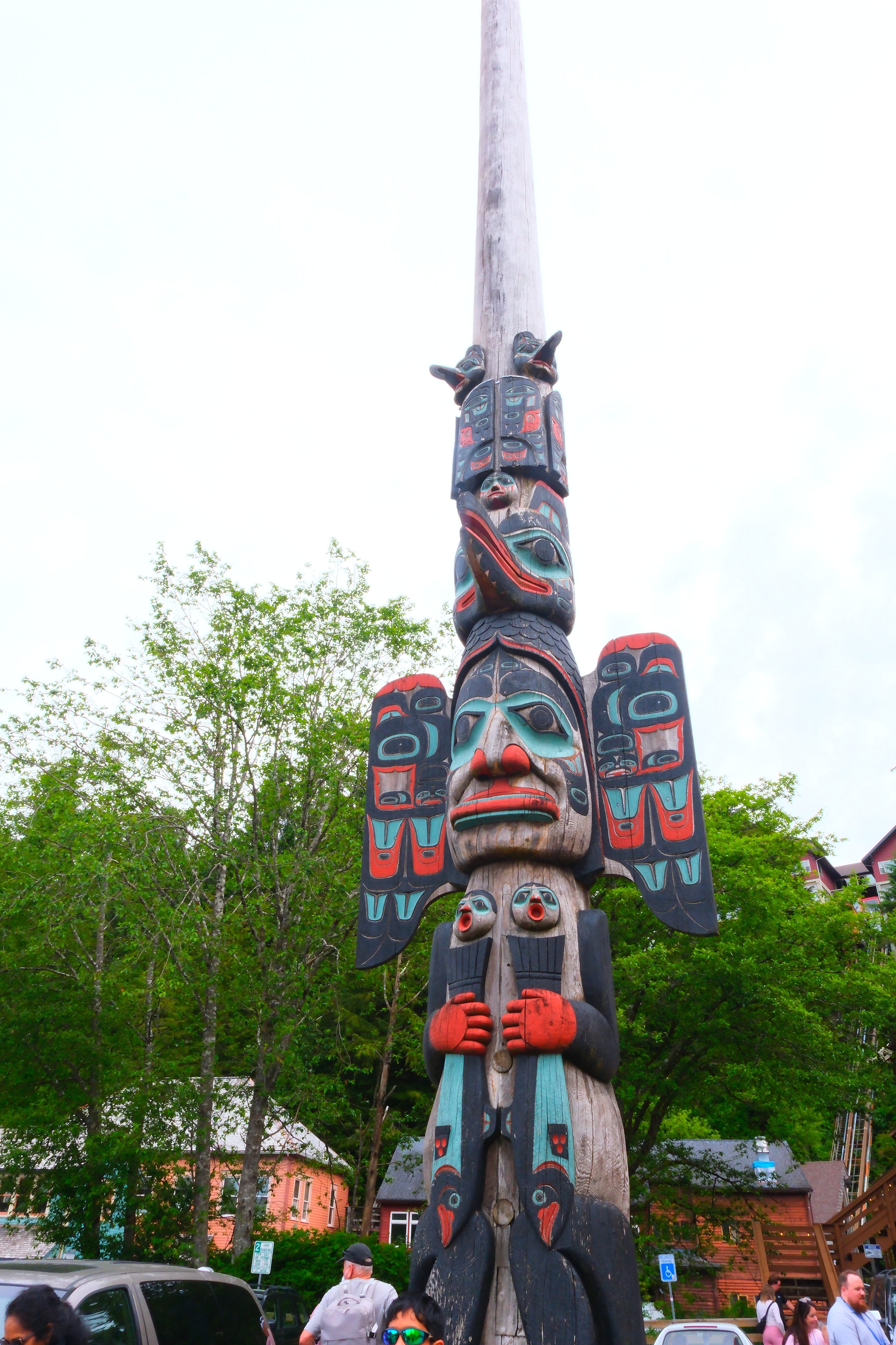 Massive totem pole with red, teal, and black Indigenous carvings rising into a white sky — downtown Ketchikan street scene behind it