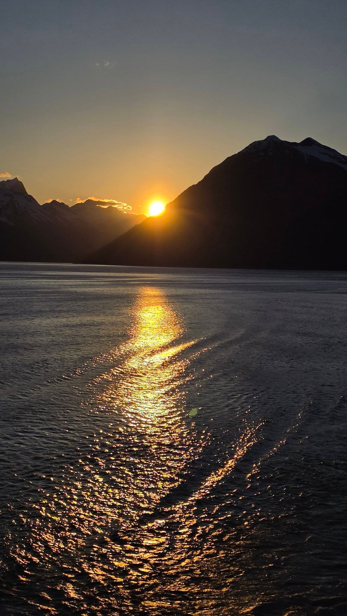 Golden sunset over the Alaska mountains departing Skagway — the sun dips between dark silhouetted peaks, its reflection stretching across the calm water of the Lynn Canal in a brilliant golden path