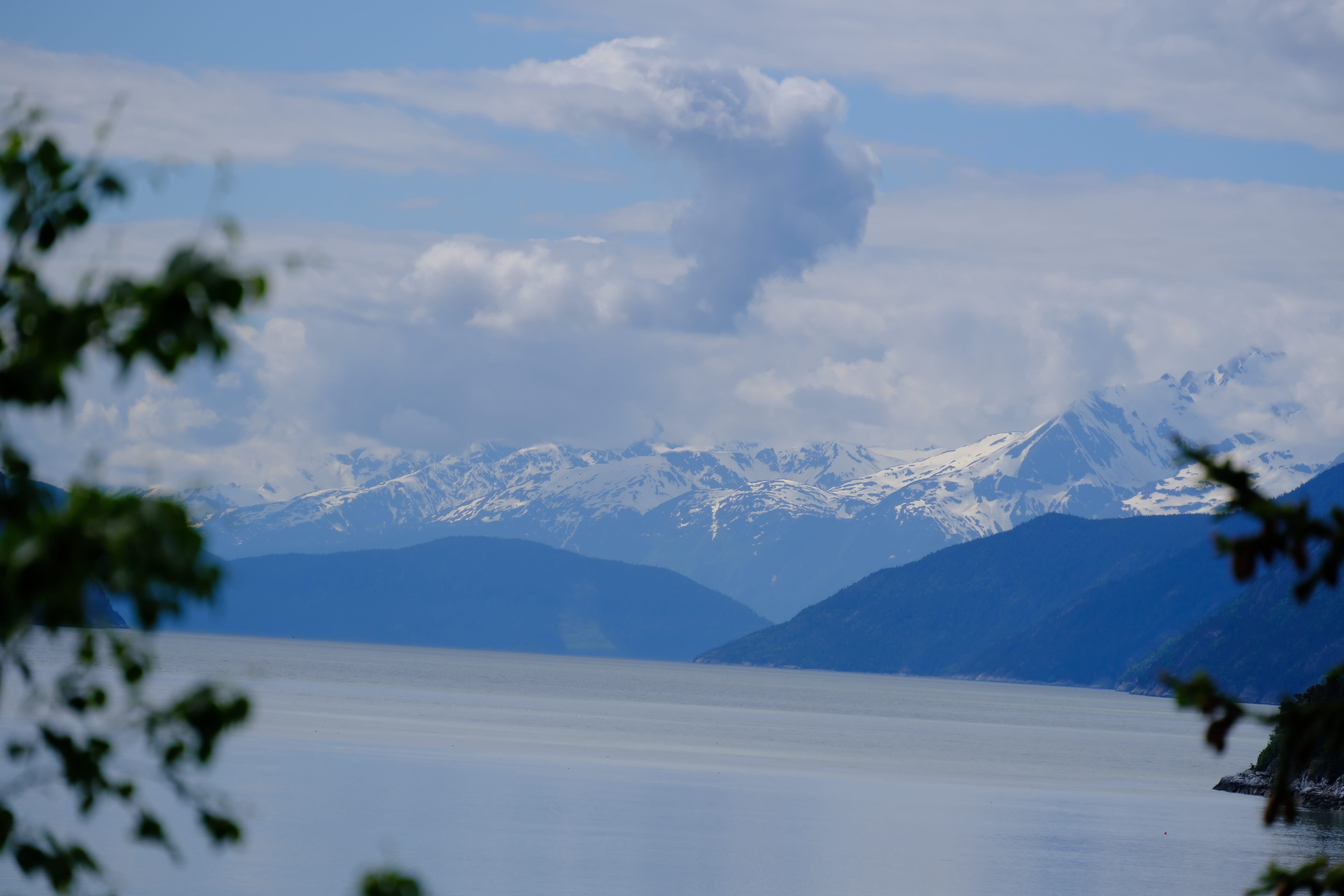 Snow-capped Alaska Range reflected in the still surface of the Lynn Canal — mountain peaks and blue sky mirrored in the glassy water, a surreal double image of the landscape