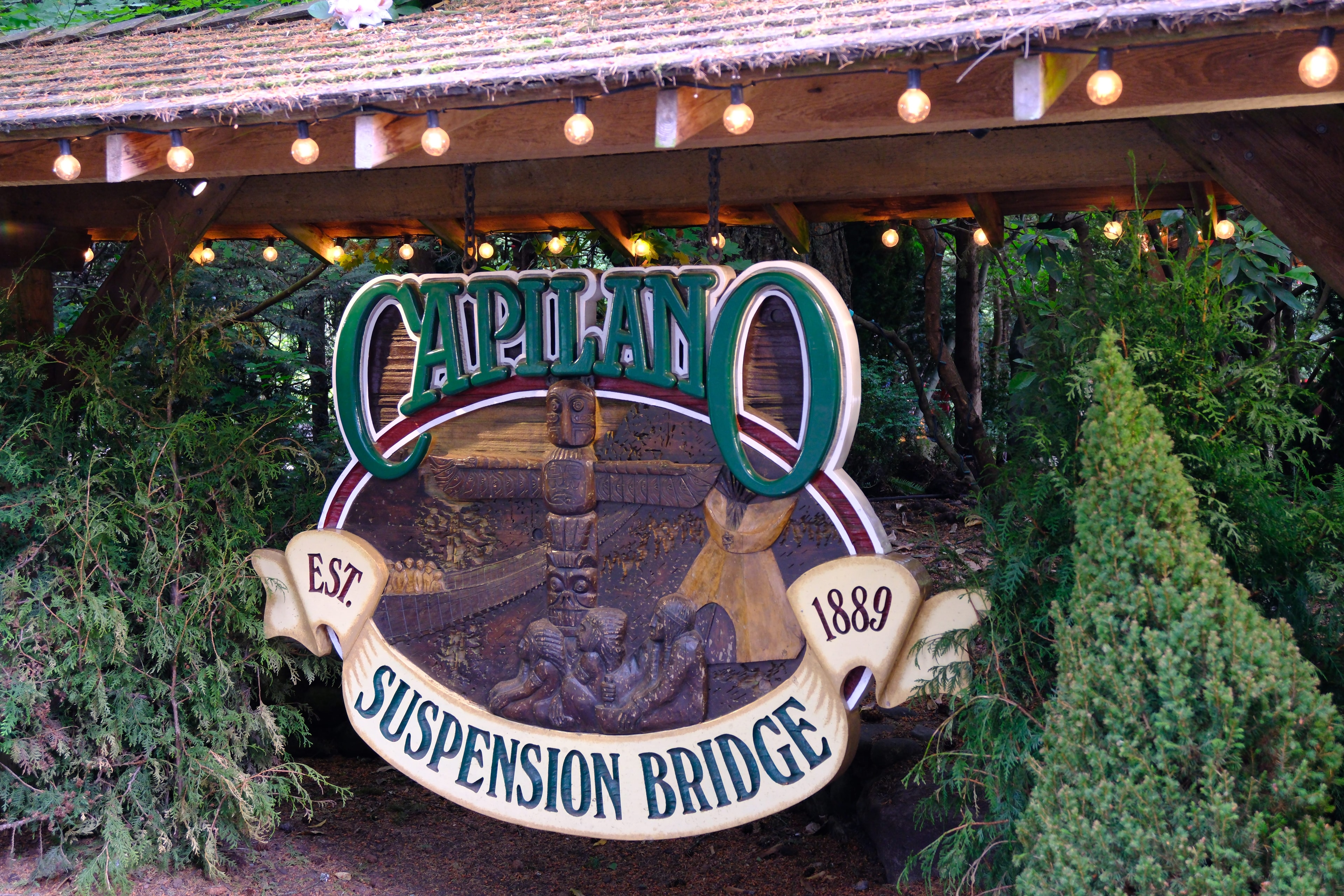 Capilano Suspension Bridge carved wooden sign — established 1889 — glowing under string lights in the forest