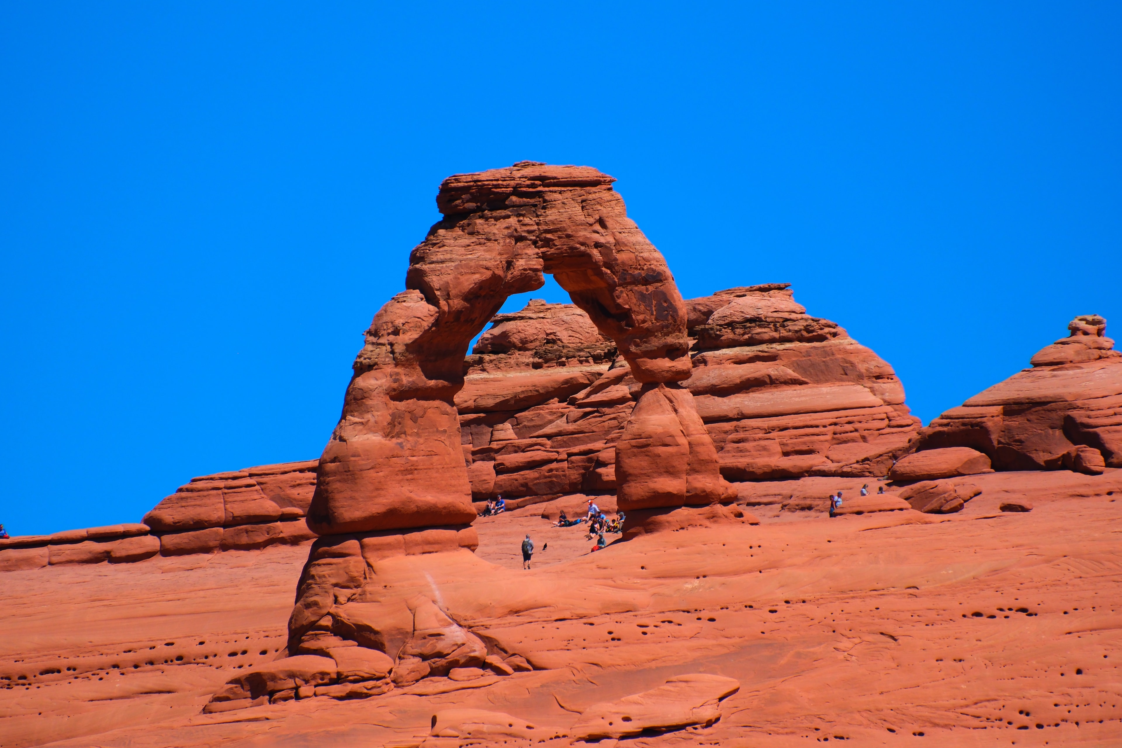 Delicate Arch classic straight-on view with visitors at the base and on the slickrock and deep blue sky at Arches National Park Utah