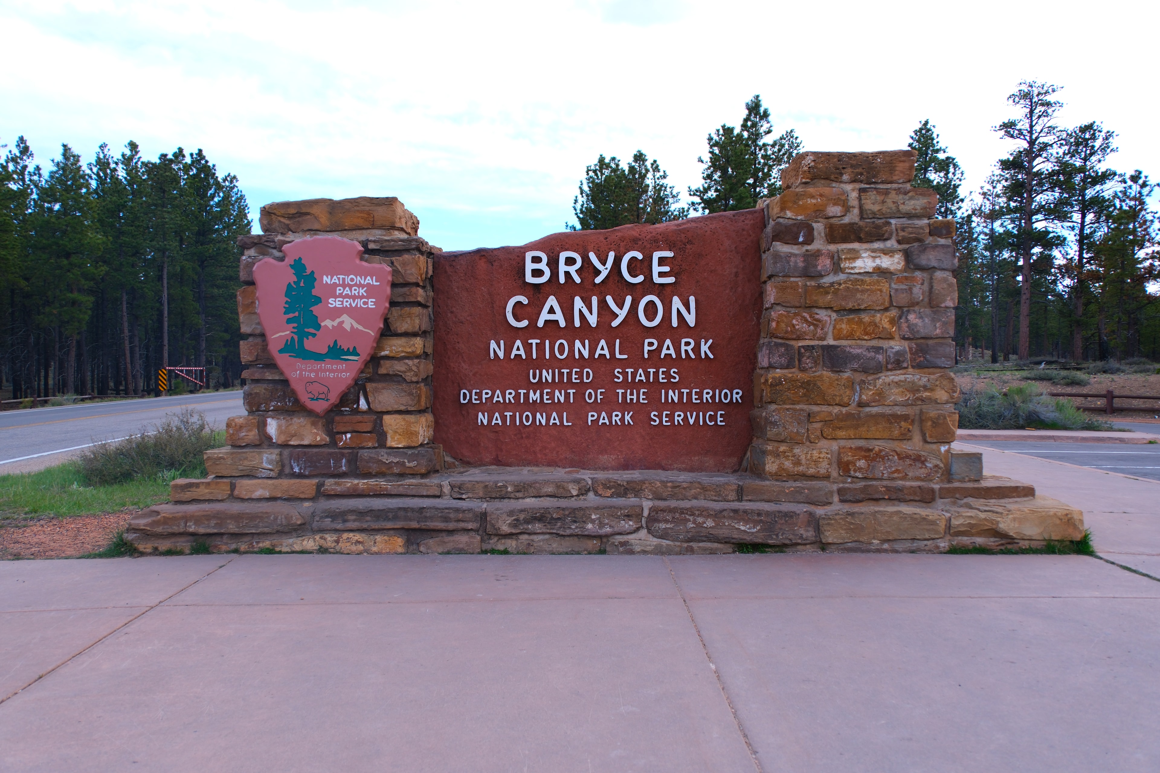 Bryce Canyon National Park entrance sign with NPS arrowhead badge and ponderosa pines behind and overcast sky Utah