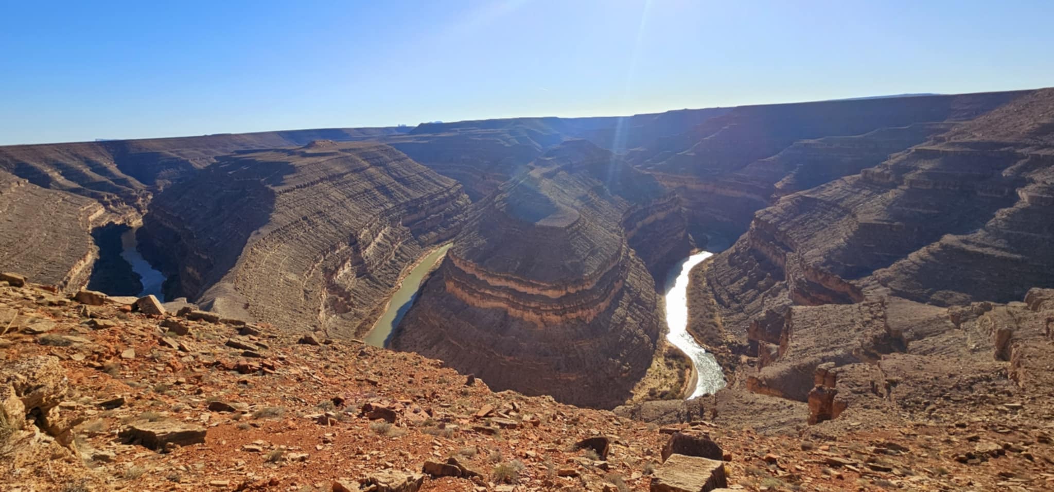 Wide view of the San Juan River horseshoe meander from the Goosenecks State Park overlook in Utah with red rock canyon walls and sun flare
