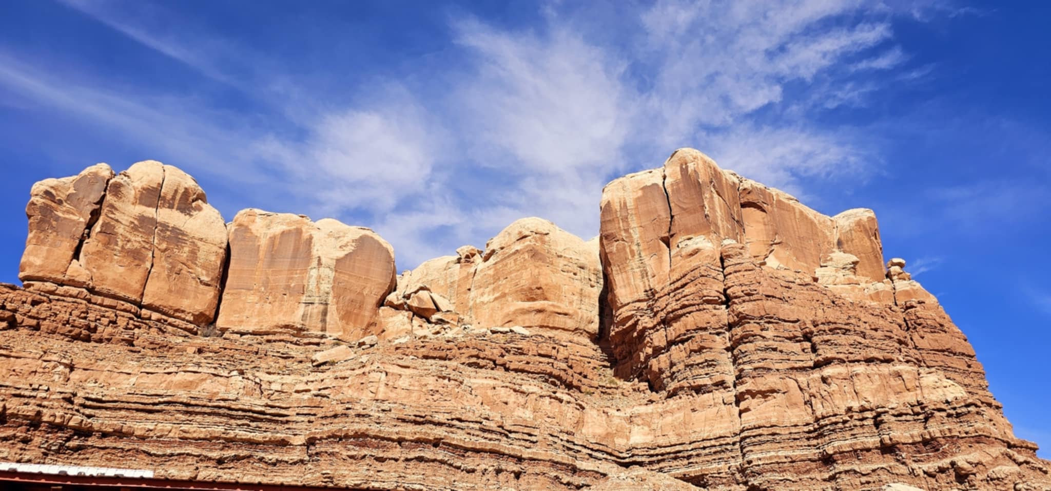 Massive layered tan and rust sandstone cliff wall with rounded cap rocks looking up against blue sky near Bluff Utah