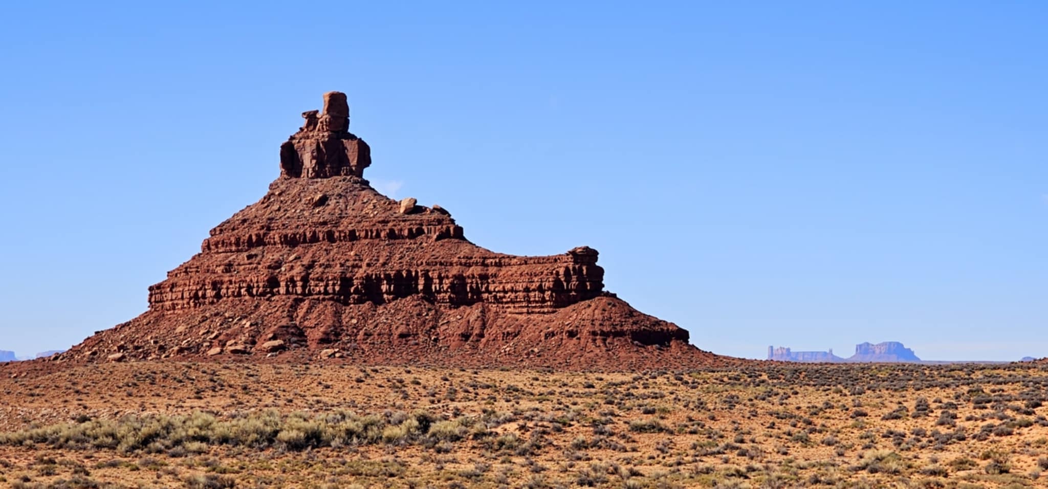 Lone pyramid-shaped sandstone butte rising from the wide open desert scrubland with Monument Valley buttes visible on the distant horizon in Valley of the Gods Utah