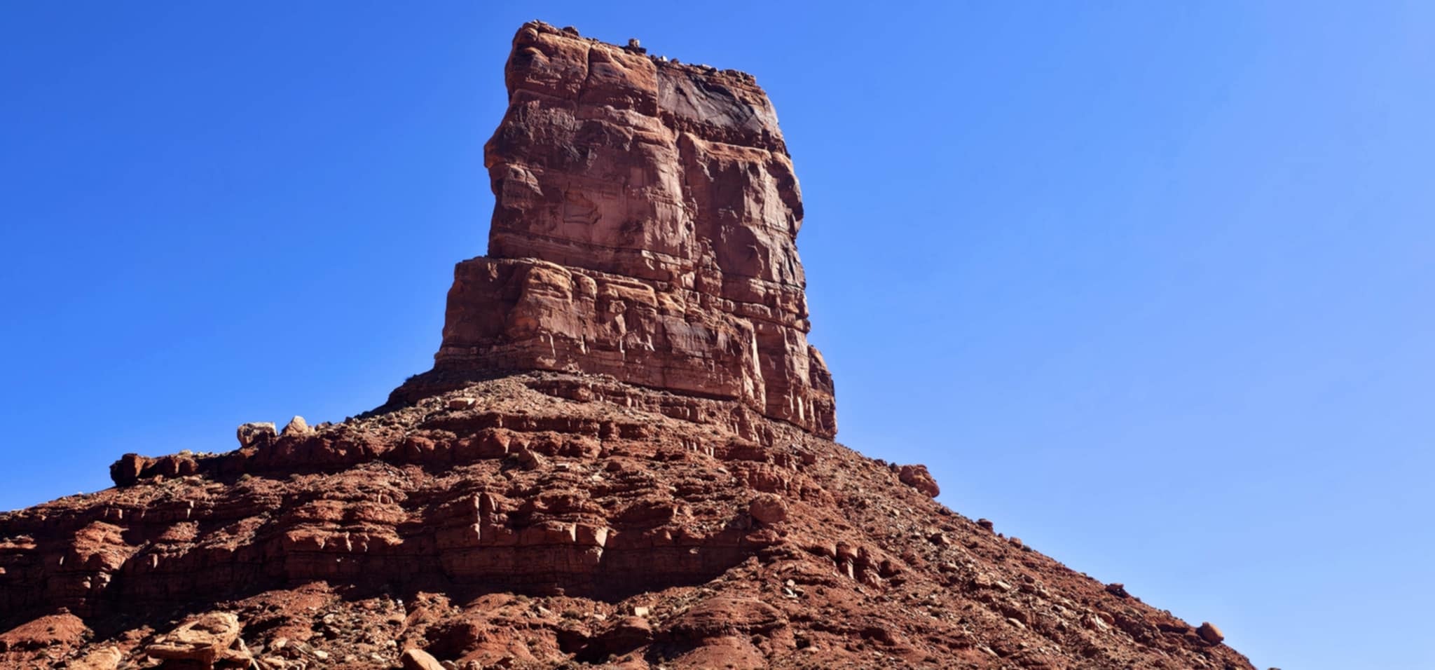 Close-up of a massive flat-topped sandstone butte with dramatic layered rock faces rising steeply against a vivid blue sky in Valley of the Gods Utah