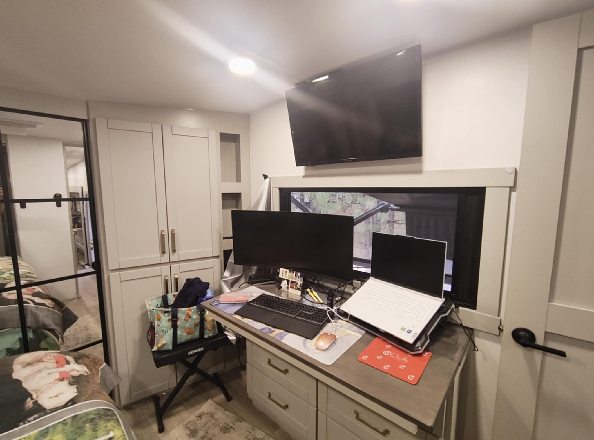 Tina's office in the Brinkley Z3400 bedroom — dresser desk with dual monitors, wall-mounted TV, and forest view through the window